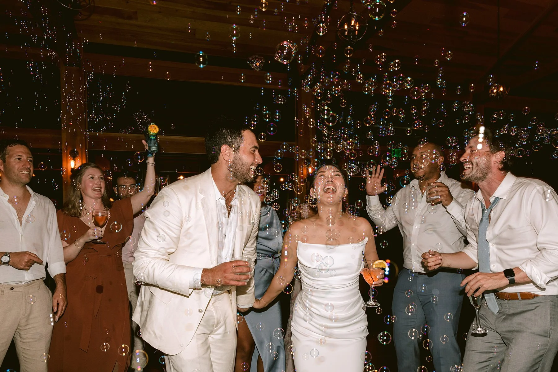A bride laughs and looks up at bubbles while her groom and guests watch at their wedding reception