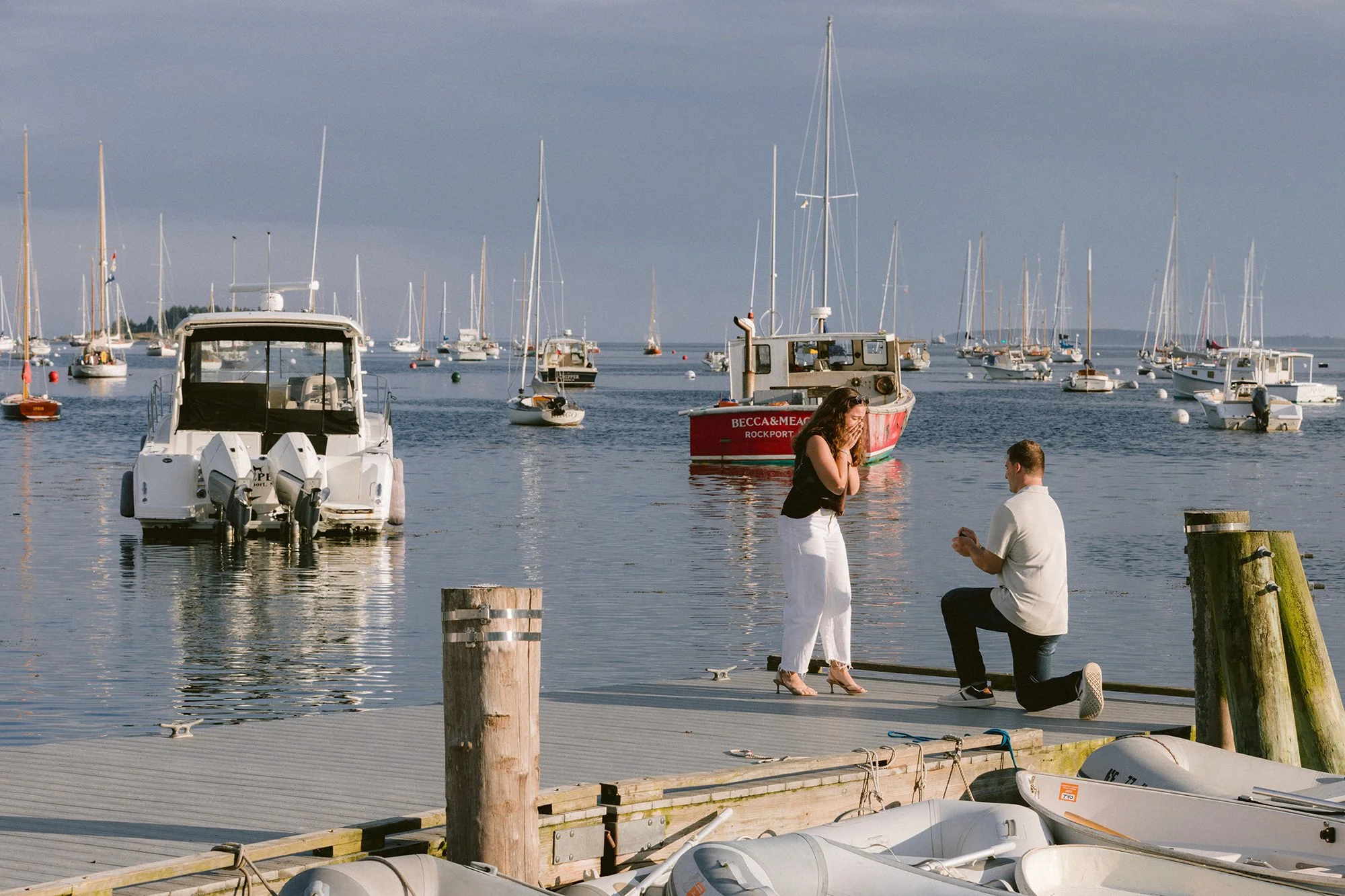 A man proposes to his girlfriend in Rockland, Maine on a dock with boats in the background