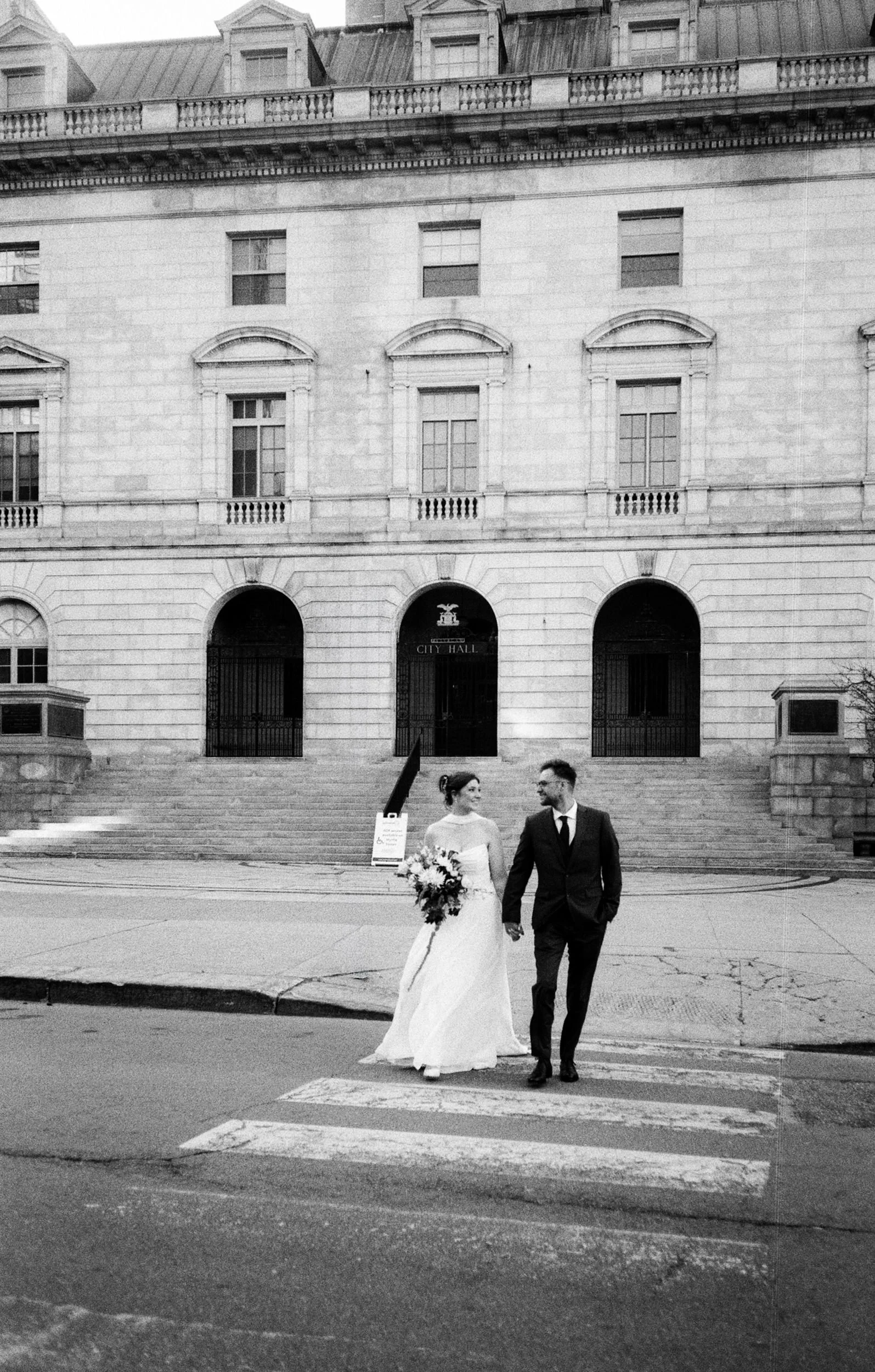 A black and white photo of a newlywed couple walking across a city street at the crosswalk in front of a large historic city hall building, with the bride holding a bouquet of flowers and the groom in a suit.