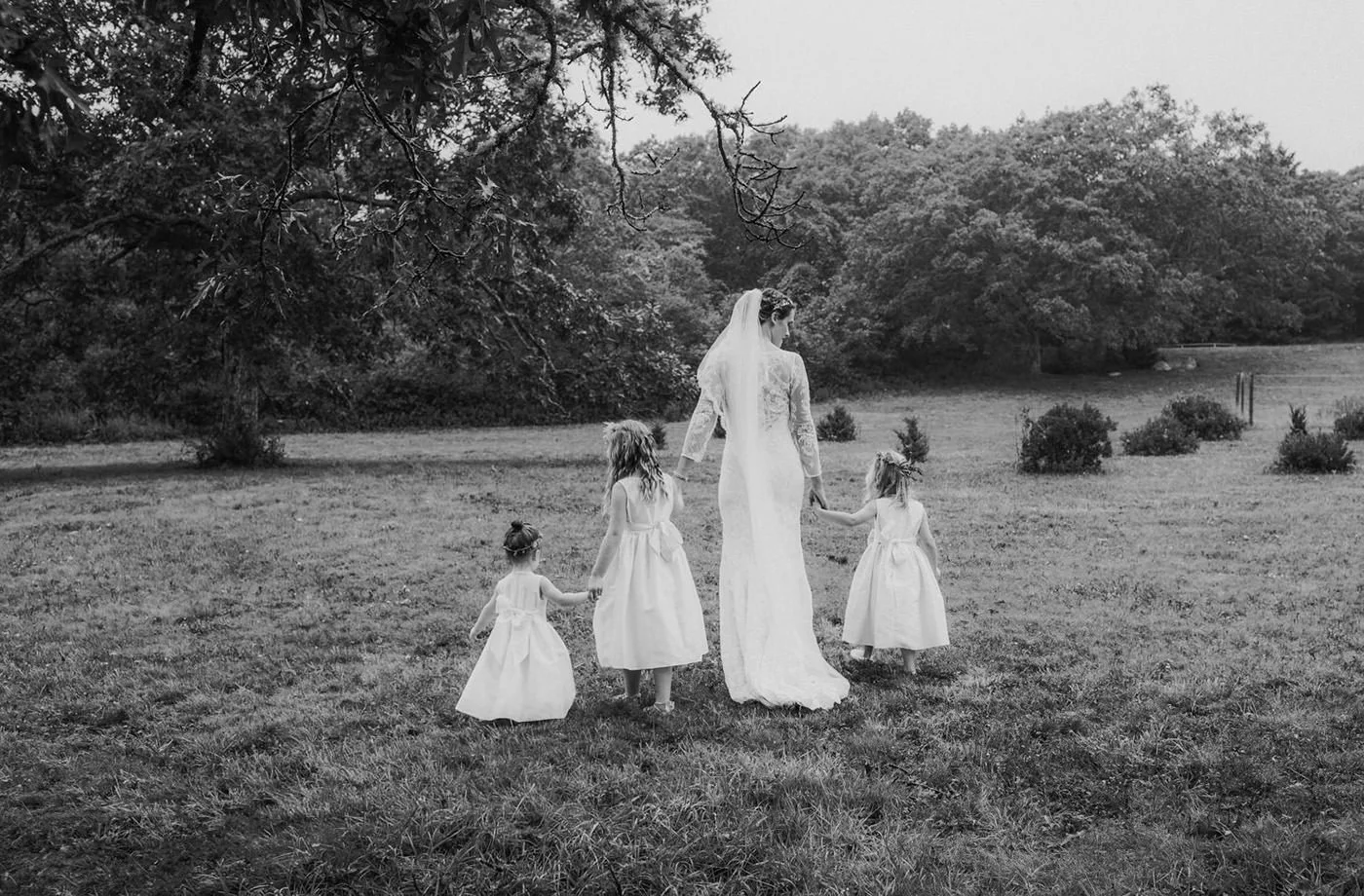 A bride walks with her 3 small flower girls, all dressed in white in a black and white photograph