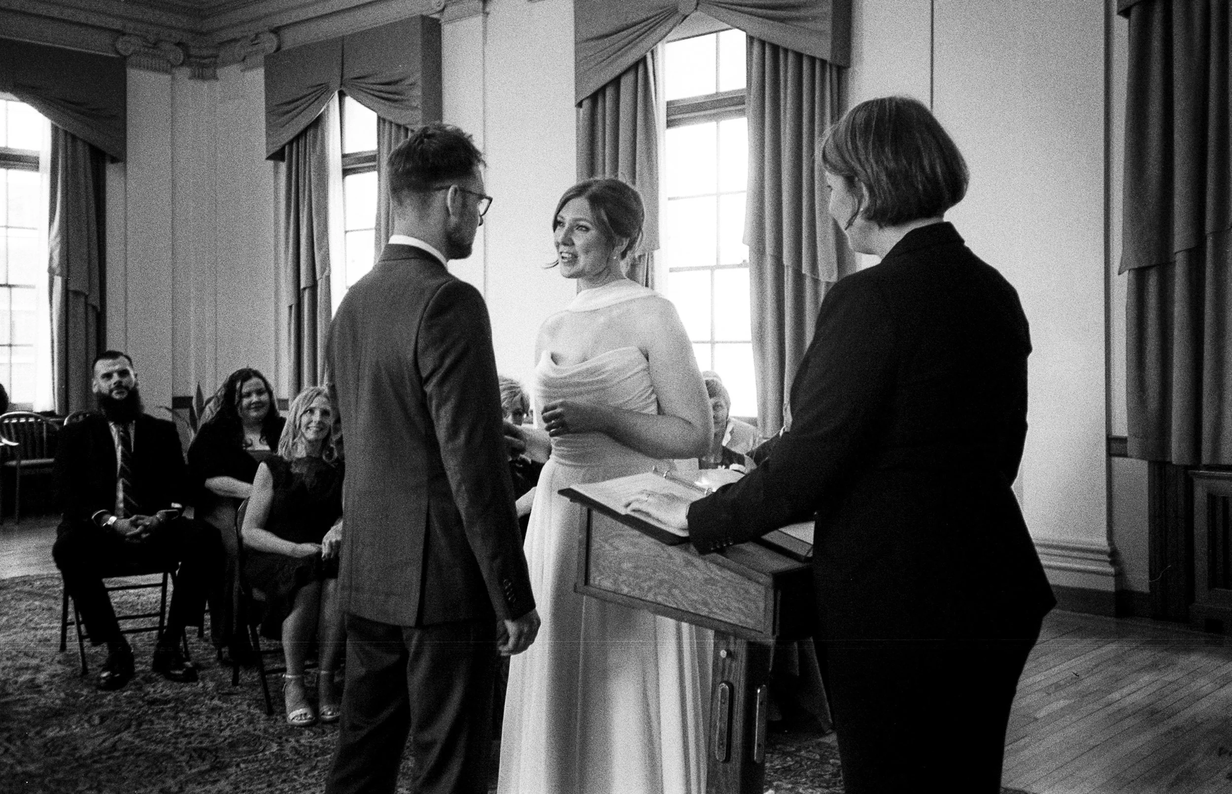 A wedding ceremony where a bride and groom stand in front of an officiant, exchanging vows, with guests seated in the background.