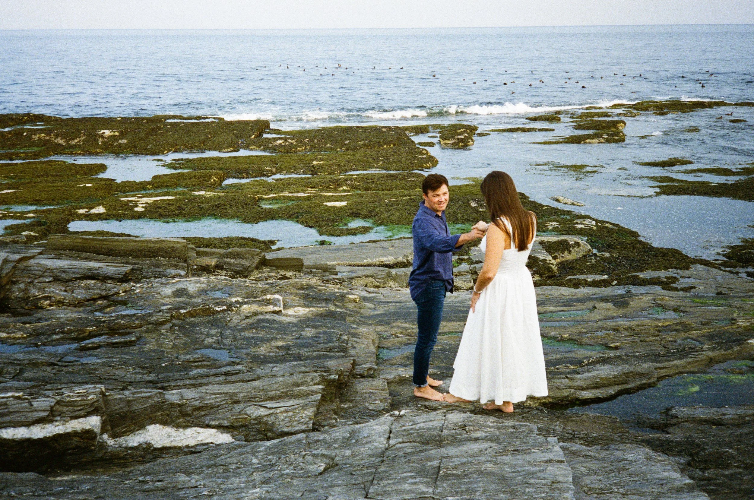 A couple stands barefoot on rocky shoreline exchanging vows, with the ocean and distant birds in the background.