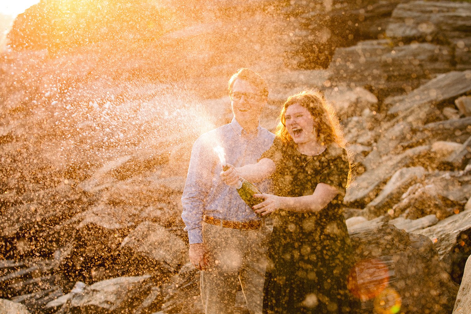 a couple who just got engaged pops a bottle of champagne at sunset in maine