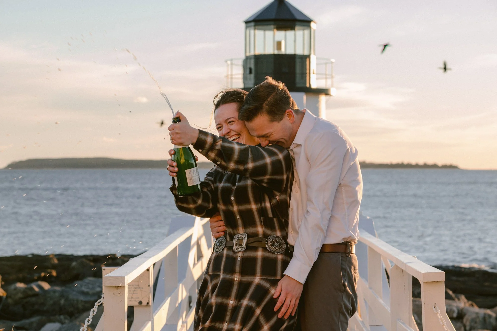 A couple sharing a joyful moment on a dock near the water, with a lighthouse and birds flying in the background during sunset.