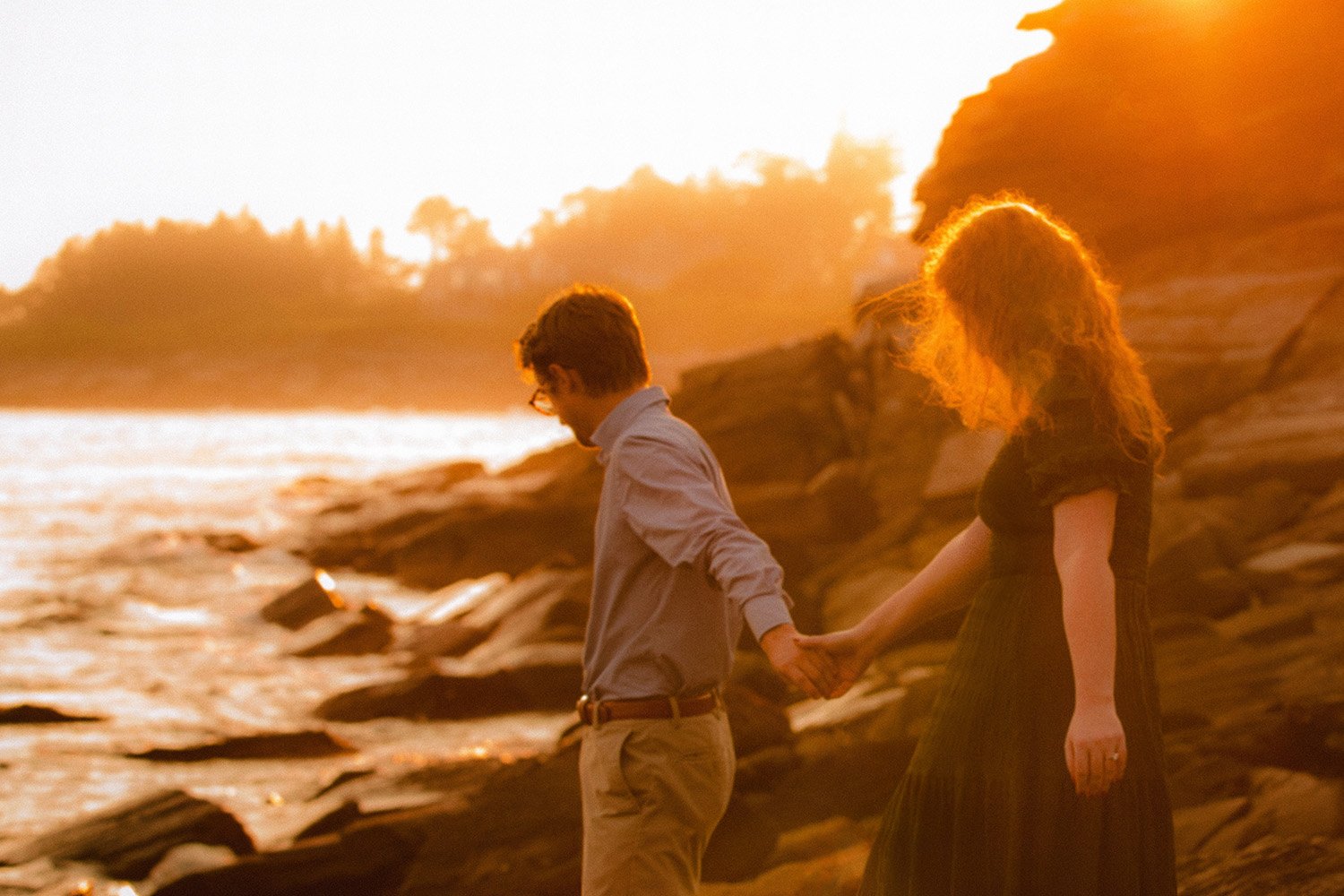 a newly engaged couple walks on the rocks in coastal maine basked in the glow of sunset light