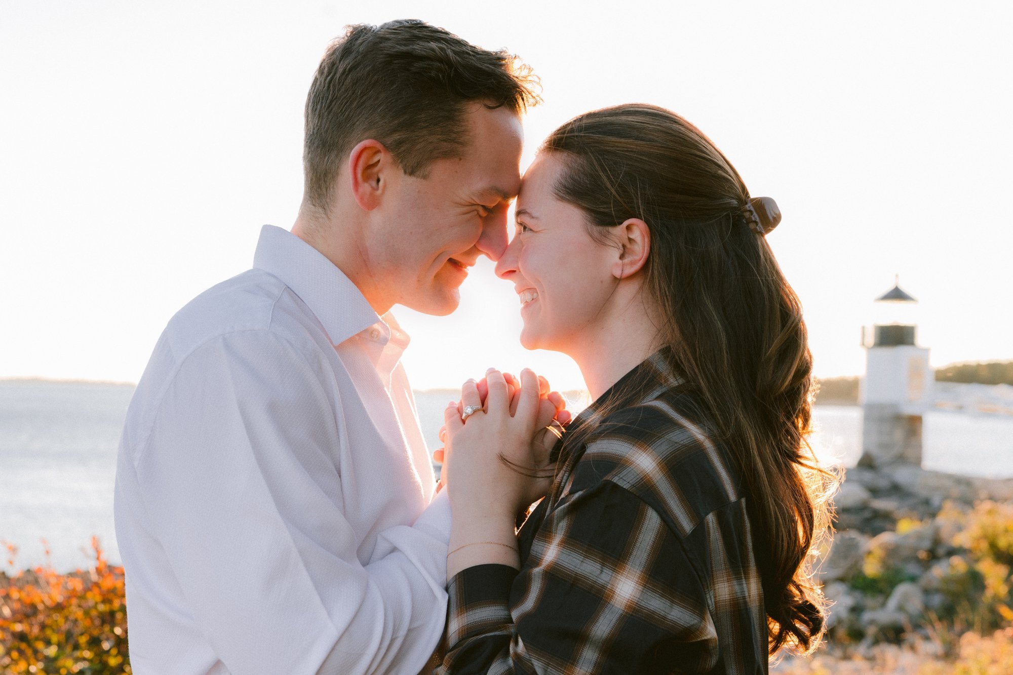 A man and woman face each other with foreheads touching, smiling and holding hands outdoors near water and a lighthouse at sunset.