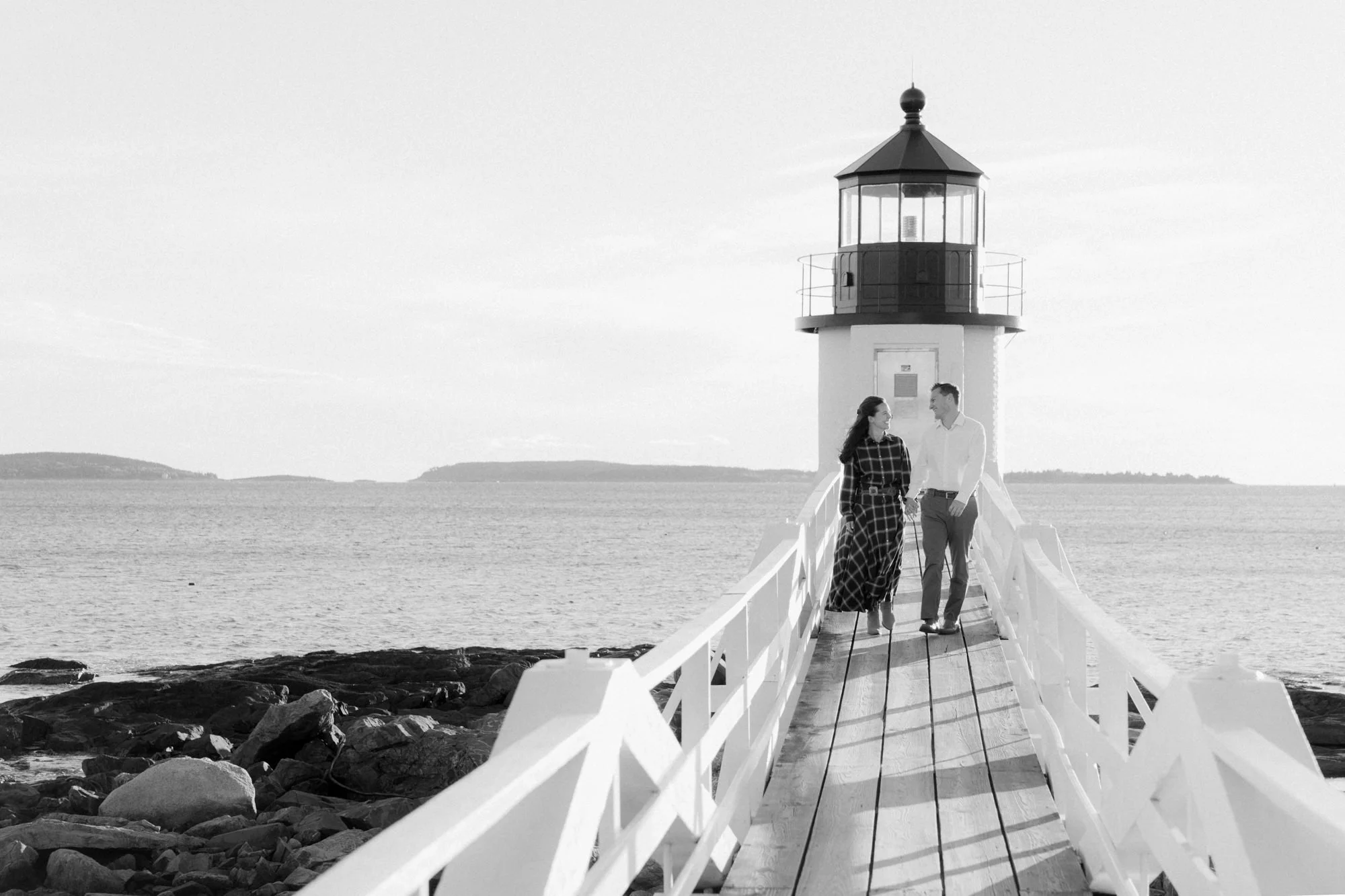 A black and white photograph of a lighthouse on a pier with a man and a woman walking together, holding hands.
