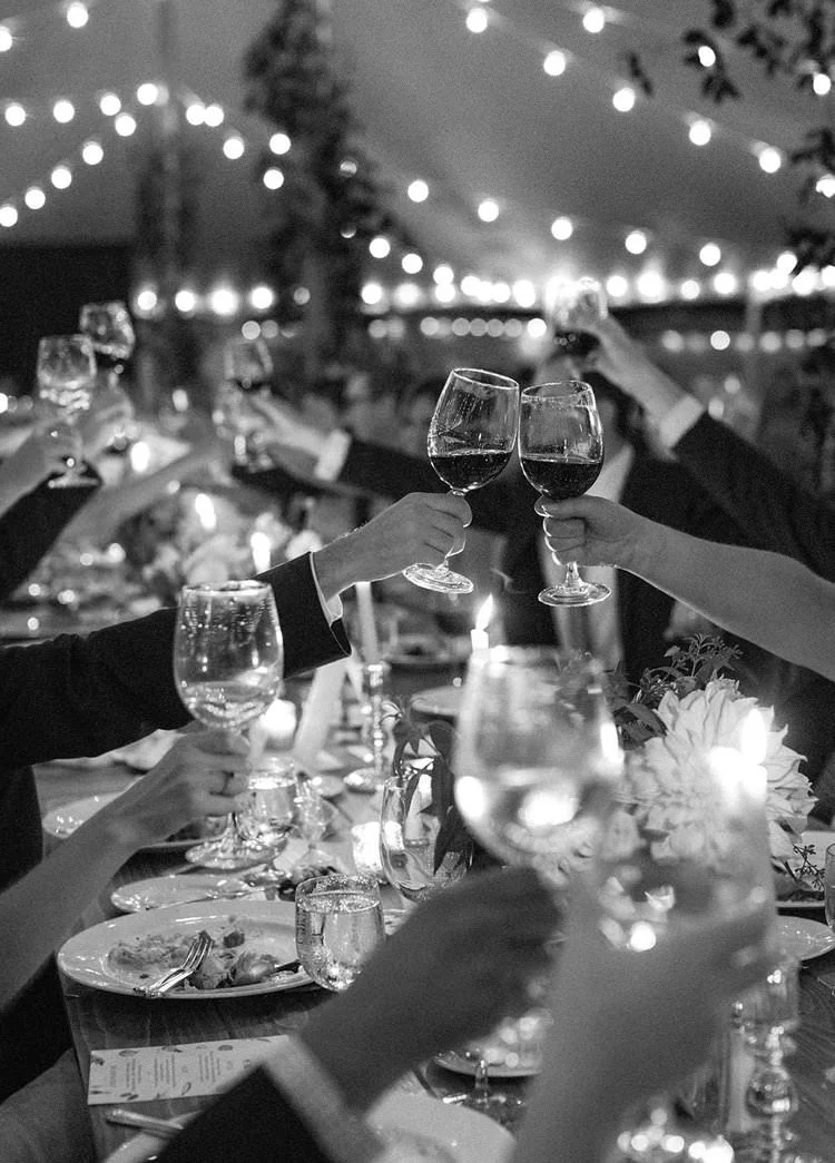 A black and white photograph of a close up of guests raising their wine glasses and saying cheers during a wedding reception by candlelight