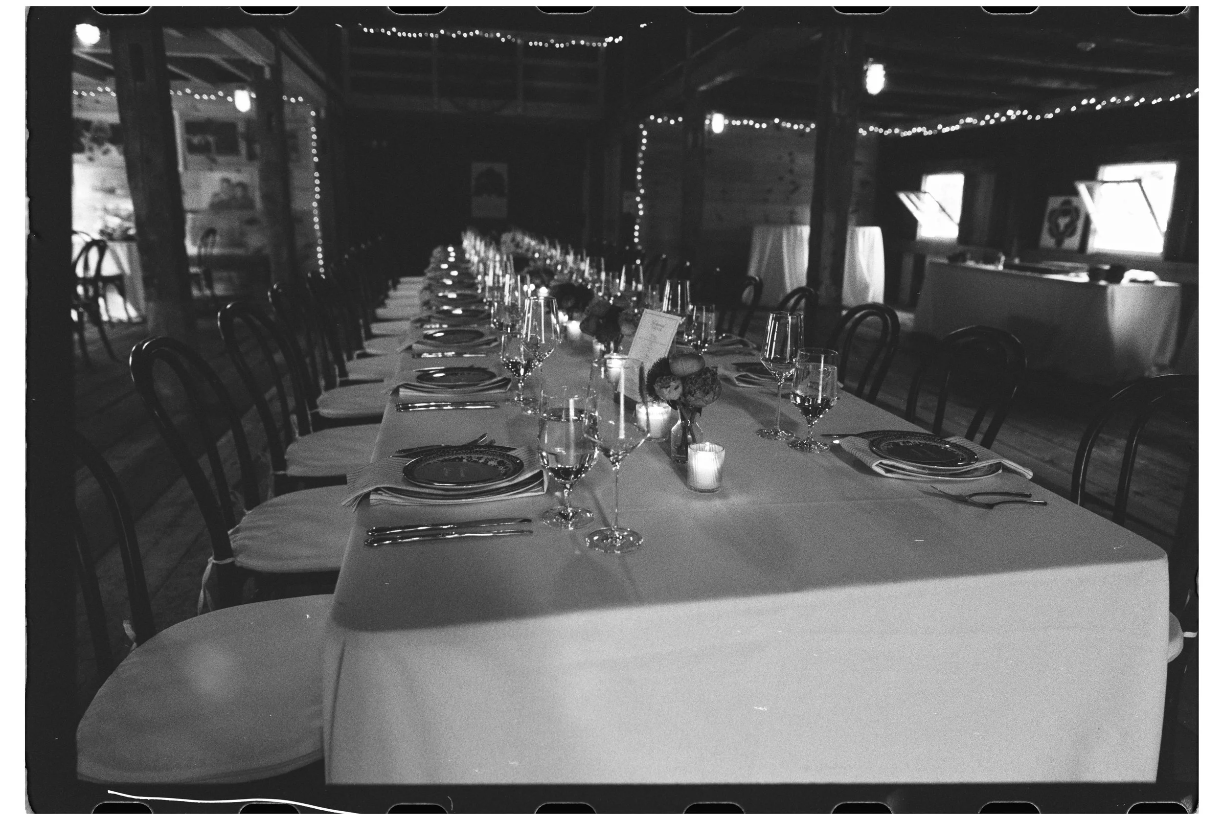 A long dining table set for a dinner event in a rustic wooden venue, decorated with wine glasses, plates, silverware, candles, and floral centerpieces, with fairy lights hanging from the ceiling.