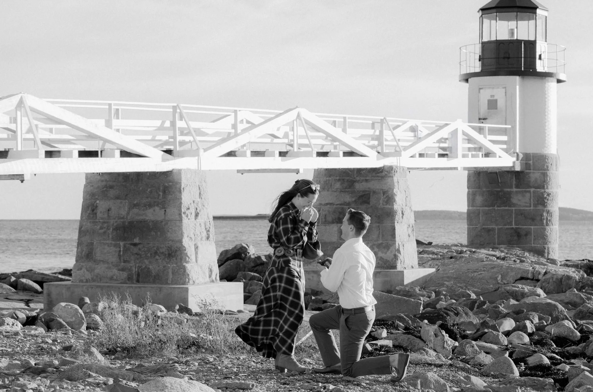 A woman appears to be proposing marriage to a man in front of a lighthouse by the water, with a rocky beach in the background.