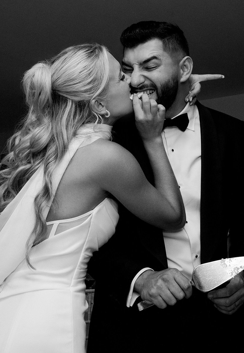 A black and white photo of a bride and groom sharing a piece of cake in a funny way