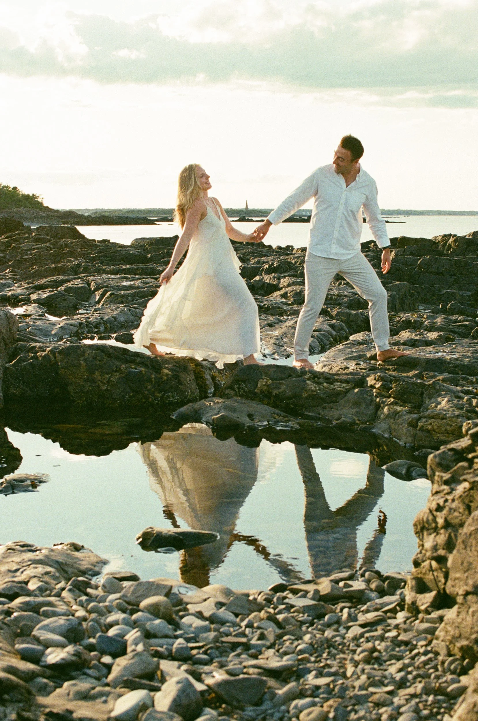 A couple holding hands and walking on a rocky beach at sunset, with their reflection visible in a small pool of water. Photographed on film in Maine