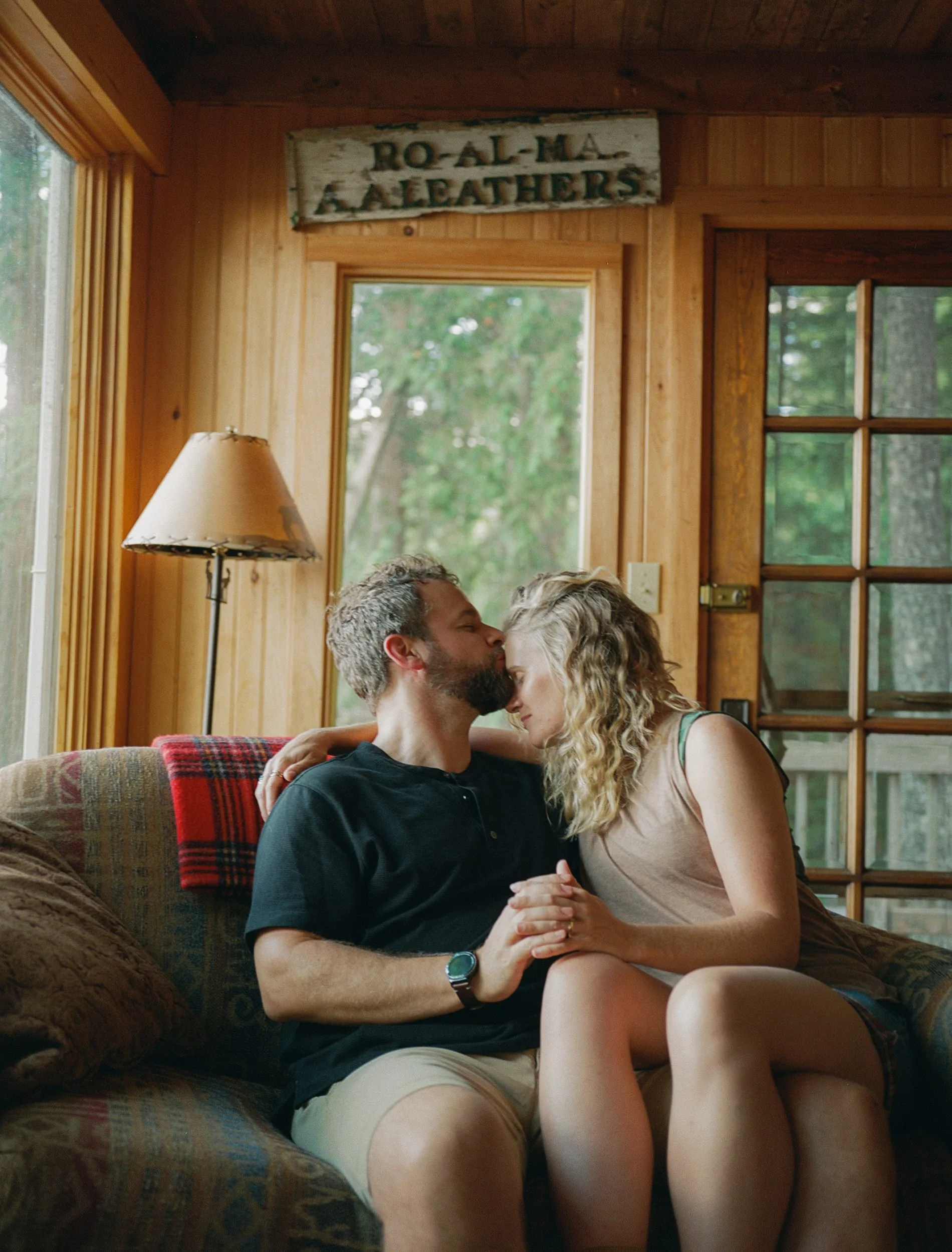 A man and woman sitting closely on a couch inside a wooden cabin, sharing an intimate moment with foreheads touching and eyes closed. Photographed on medium format, color film