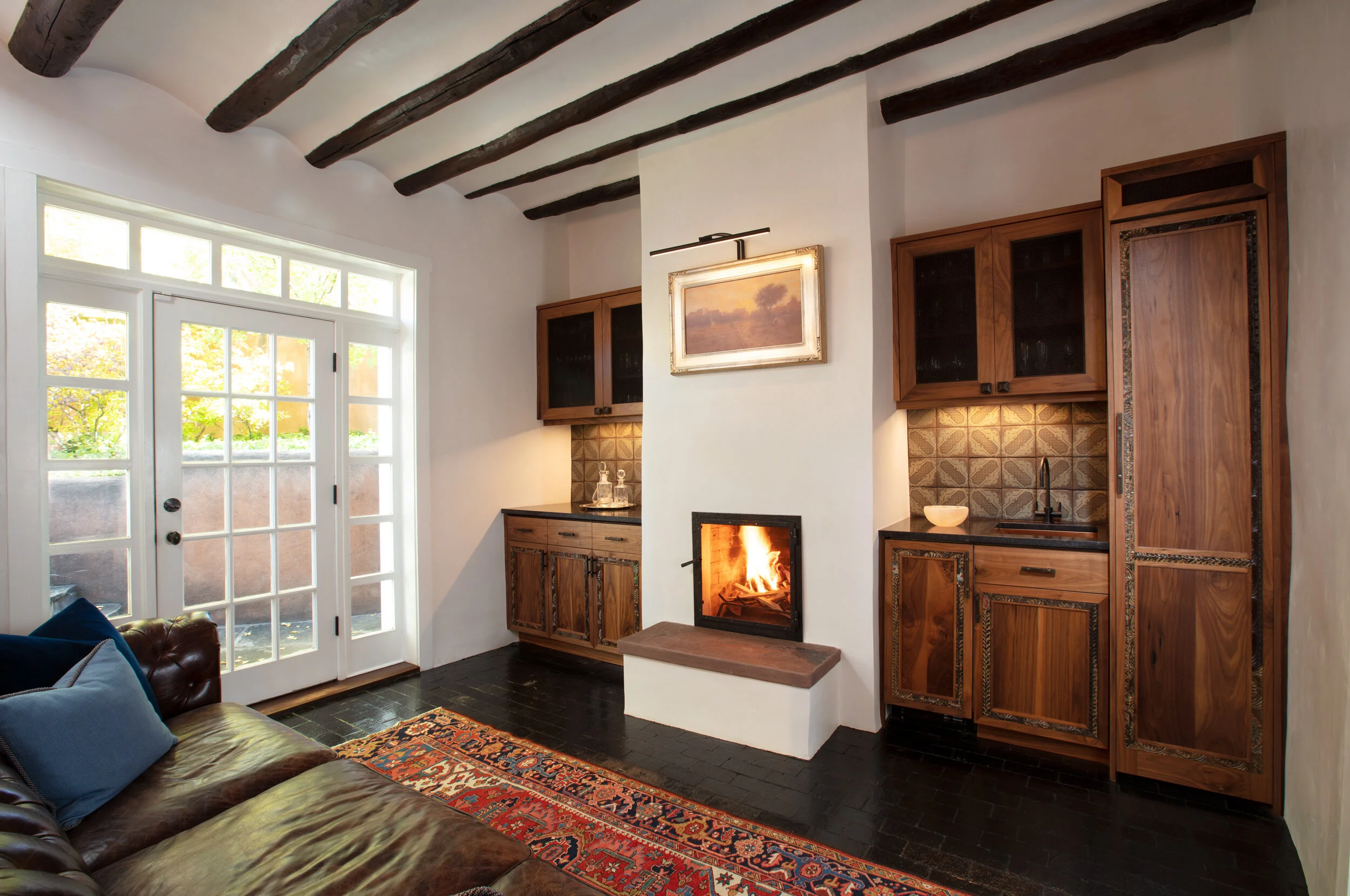 A cozy living room with a white wall and exposed dark wooden beams on the ceiling. There is a fireplace with a fire burning built into the wall, flanked by wooden cabinets. A painting hangs above the fireplace. To the left, a glass-paned door leads o