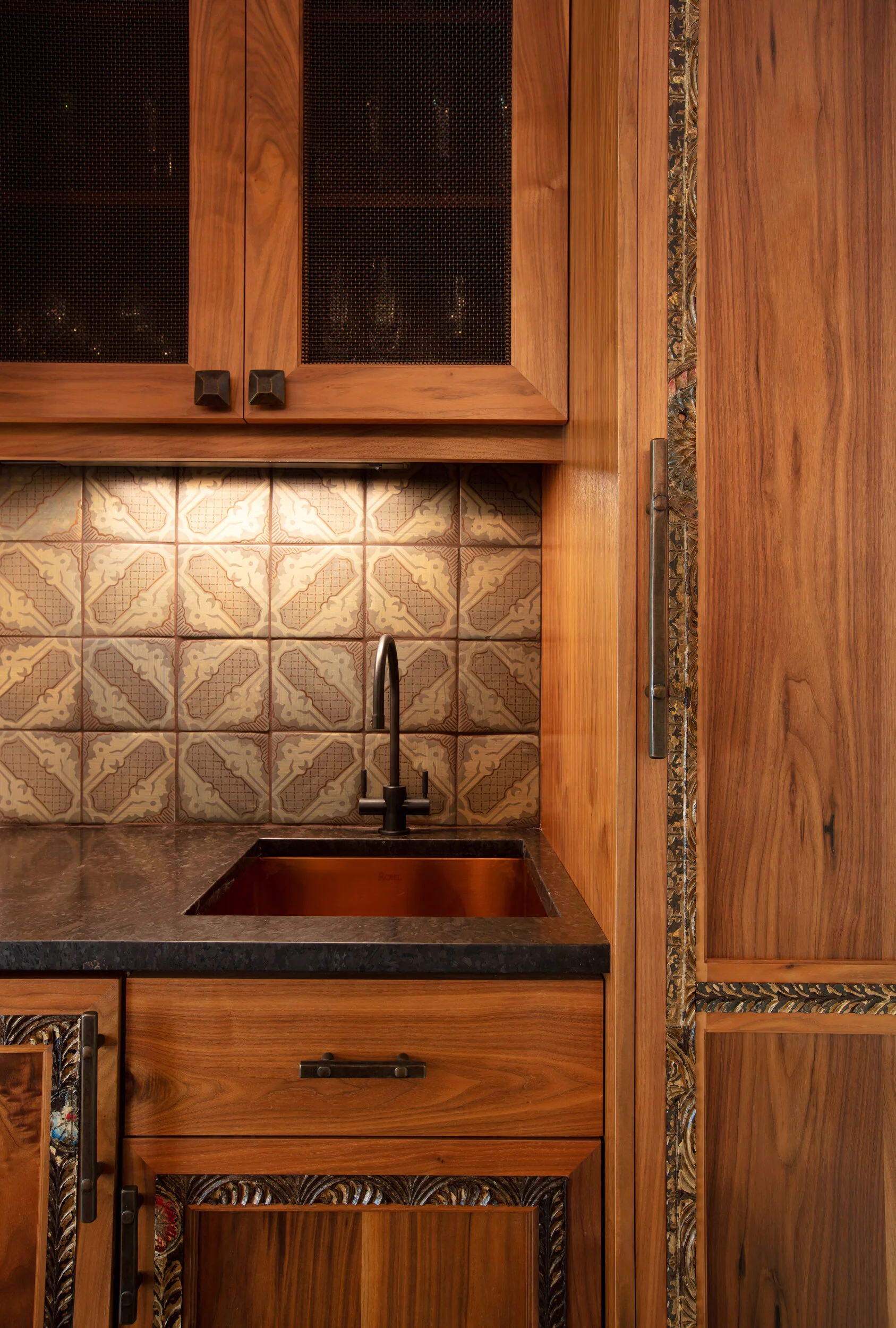 Close-up view of a rustic kitchen corner with a dark countertop, wood cabinets, decorative tiles behind a sink, and a black faucet.