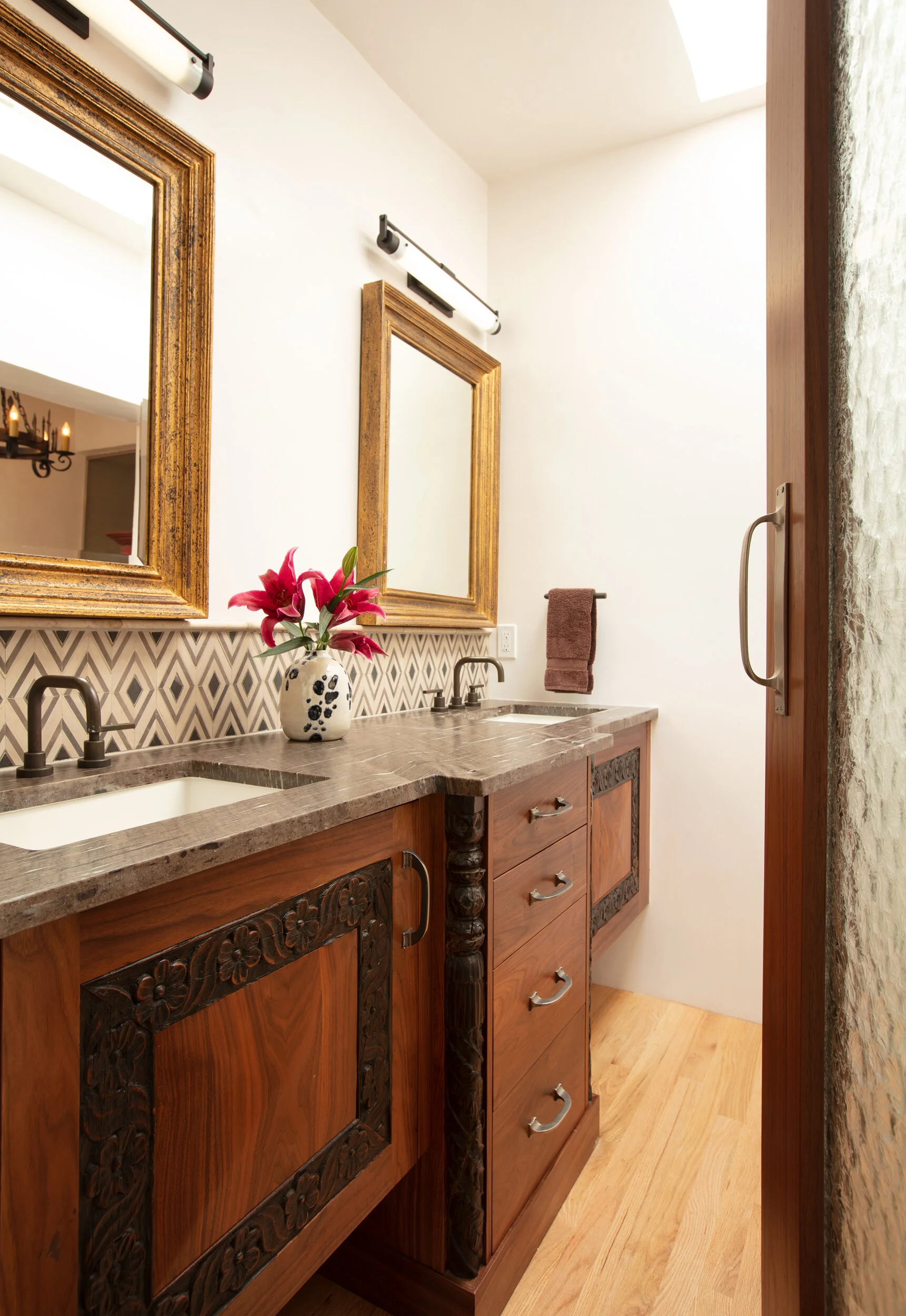 A bathroom vanity with two sinks, a patterned backsplash, a vase with pink lilies, and two wooden mirrors above the sinks.