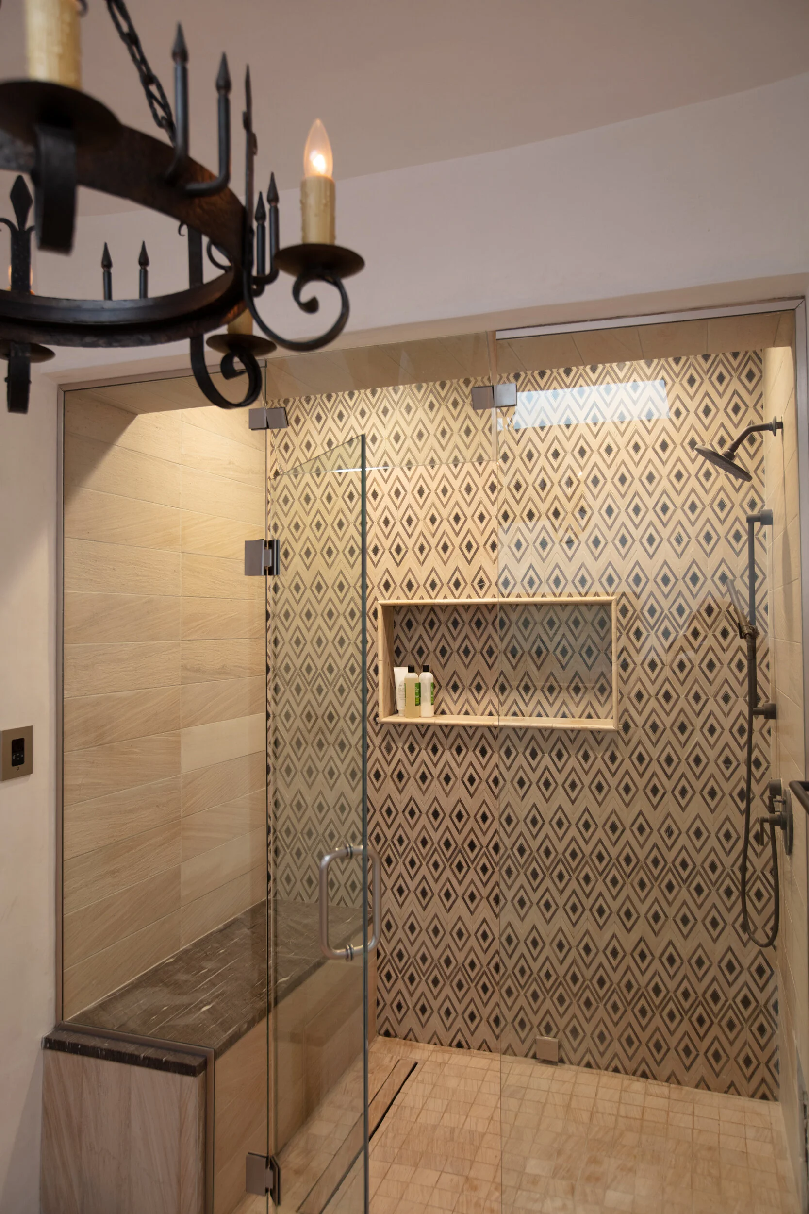 Walk-in shower with patterned tile wall, glass door, built-in shelf with toiletries, and a showerhead.