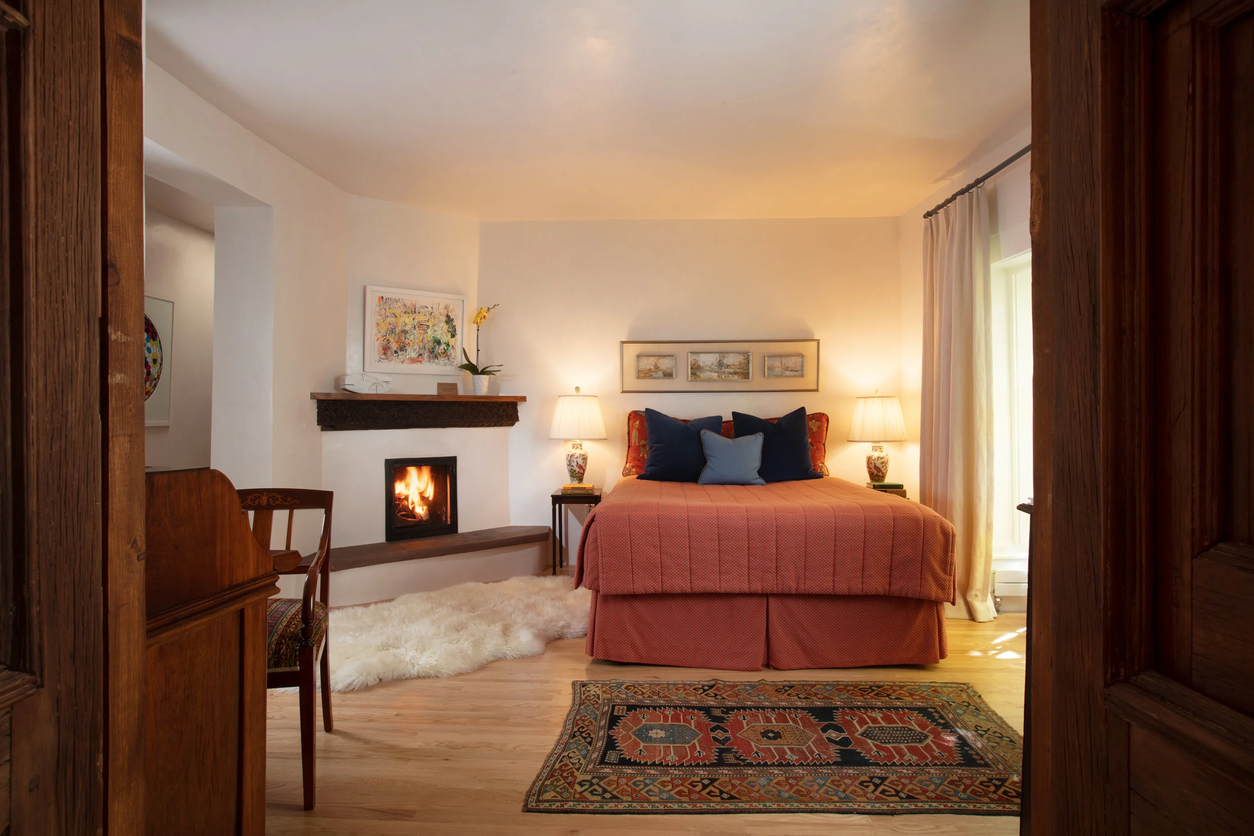 Cozy bedroom with a bed, colorful pillows, a fireplace, side tables with lamps, artwork on the wall, a rug, and curtains by the window.