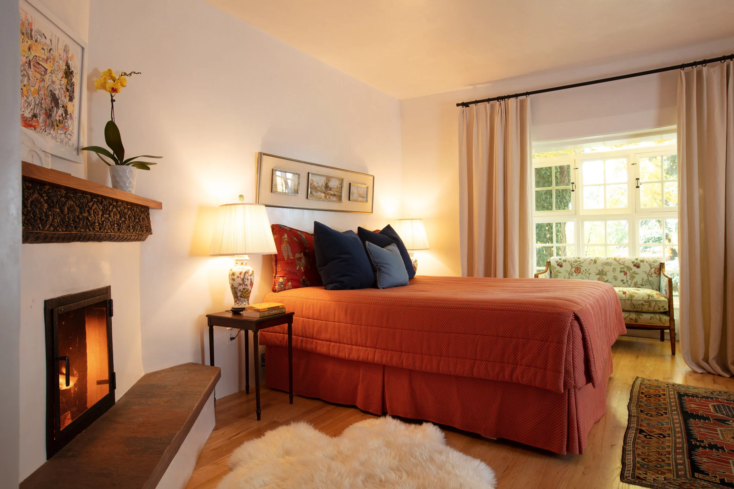 A cozy bedroom with a red bedspread, dark blue pillows, and a small nightstand with a lamp. There is a window with beige curtains and a floral sofa near the window. A fireplace with a decorative mantel is on the left, with a potted yellow orchid on t