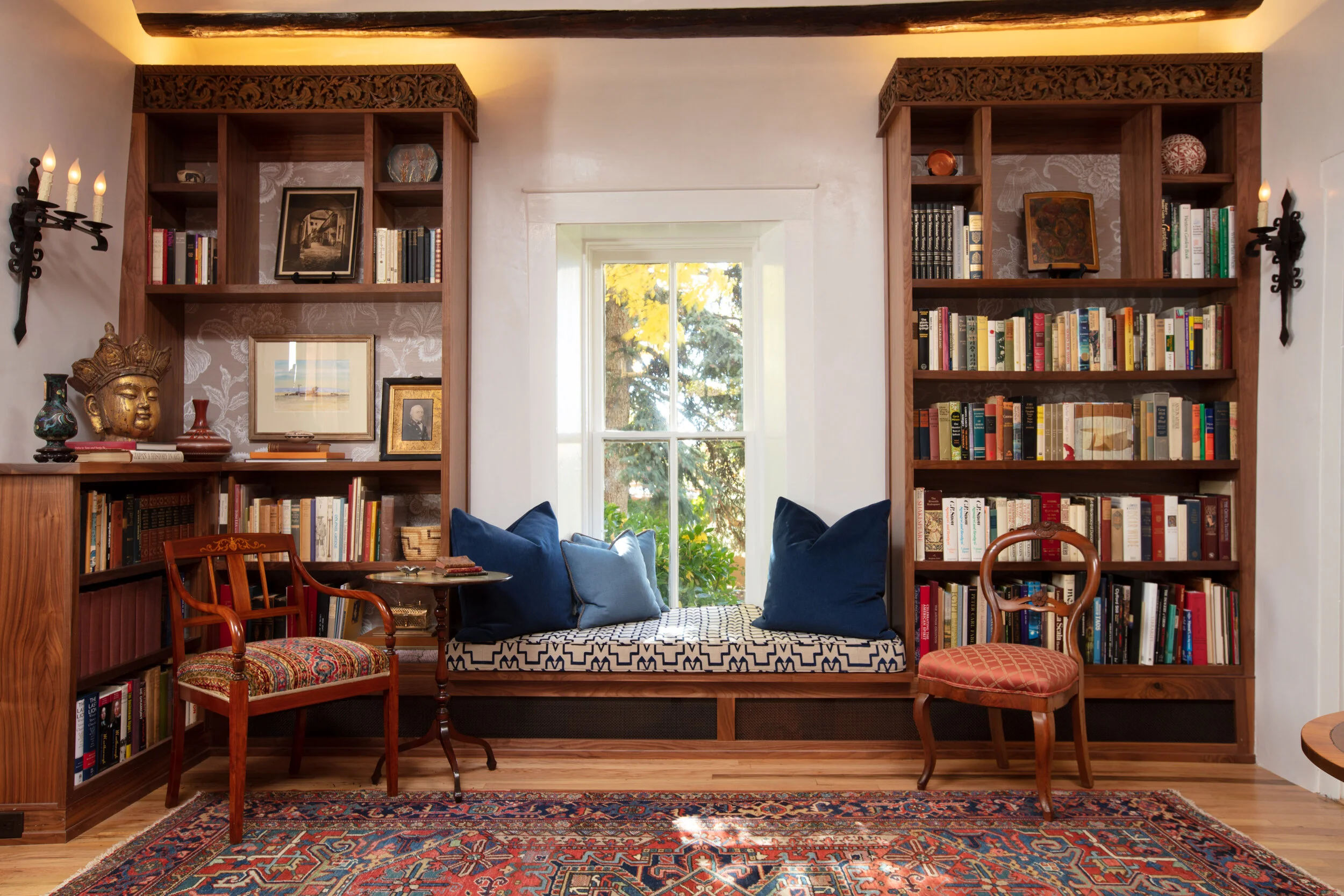 Living room with built-in bookshelves on either side of a window seat. The shelves contain books, art, and decorative objects. The window seat has three large blue pillows and overlooks trees outside. There are two vintage chairs with patterned cushi