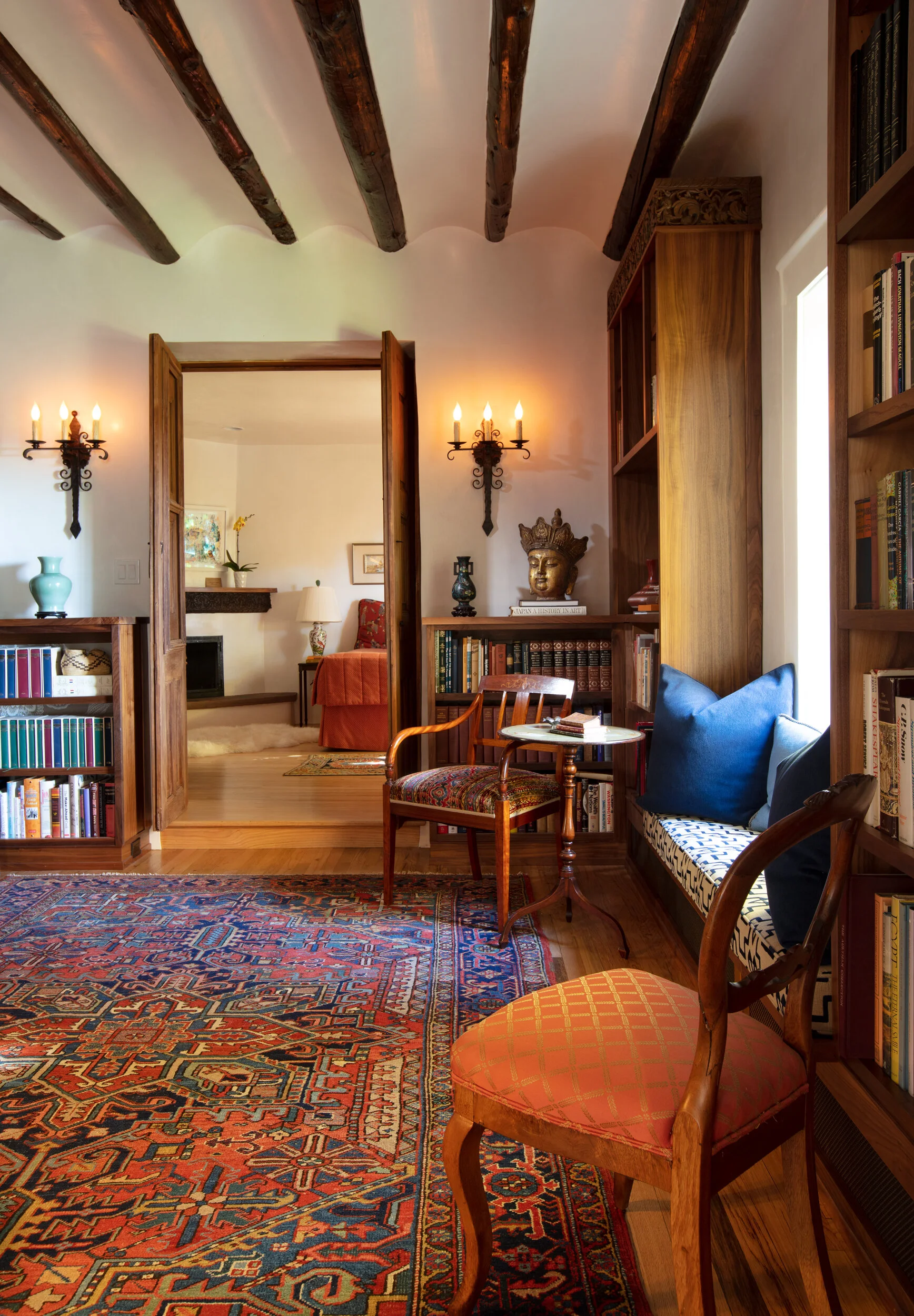 A cozy reading nook in a house with wooden beams on the ceiling, bookshelves filled with books, a small table, and vintage-style chairs with cushions, illuminated by wall sconces.