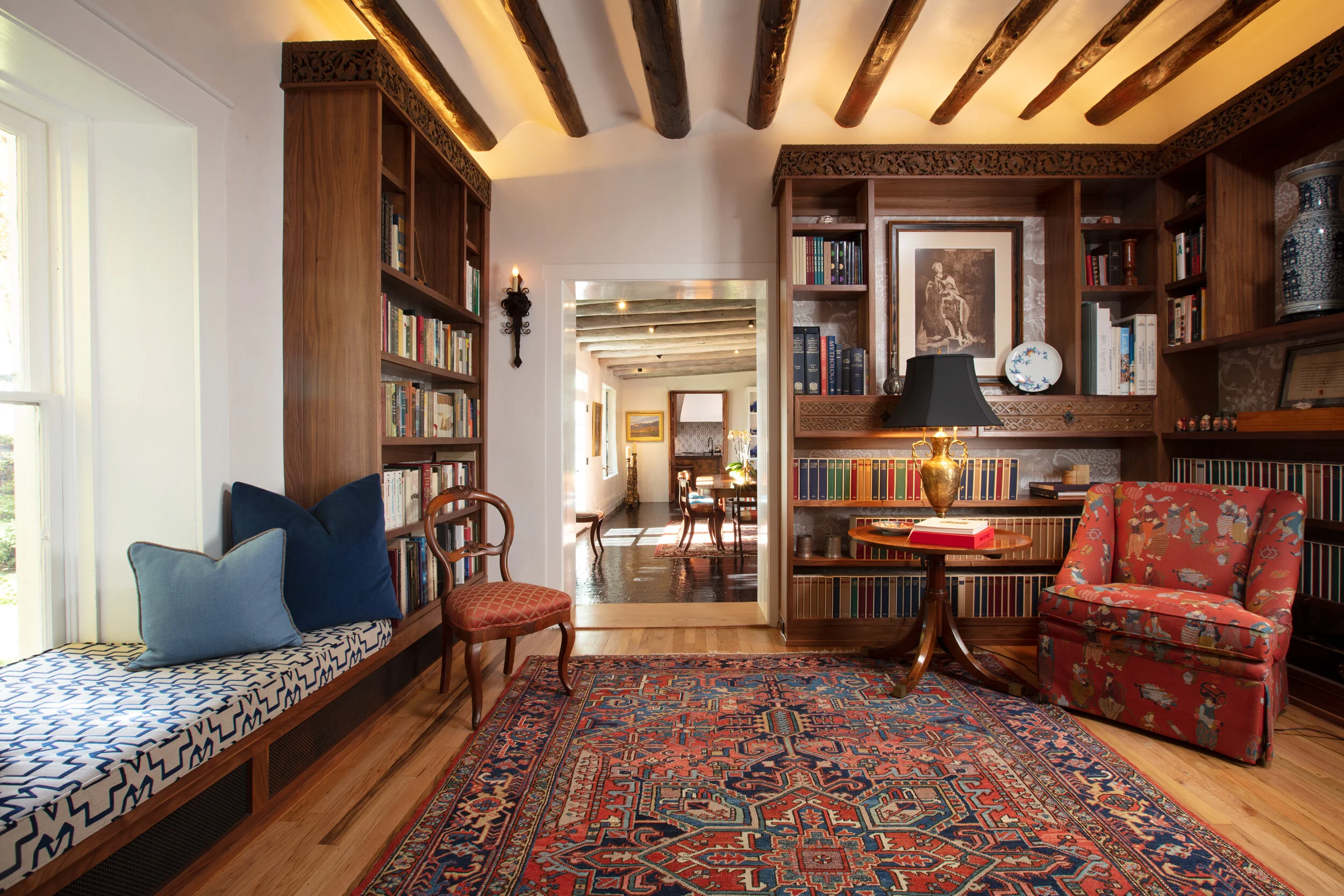 A cozy living room with hardwood flooring, built-in bookshelves filled with books, a colorful Persian rug, and various chairs and decorative items. There is a window seat with pillows, a patterned red armchair, an ornate wooden chair, a side table wi