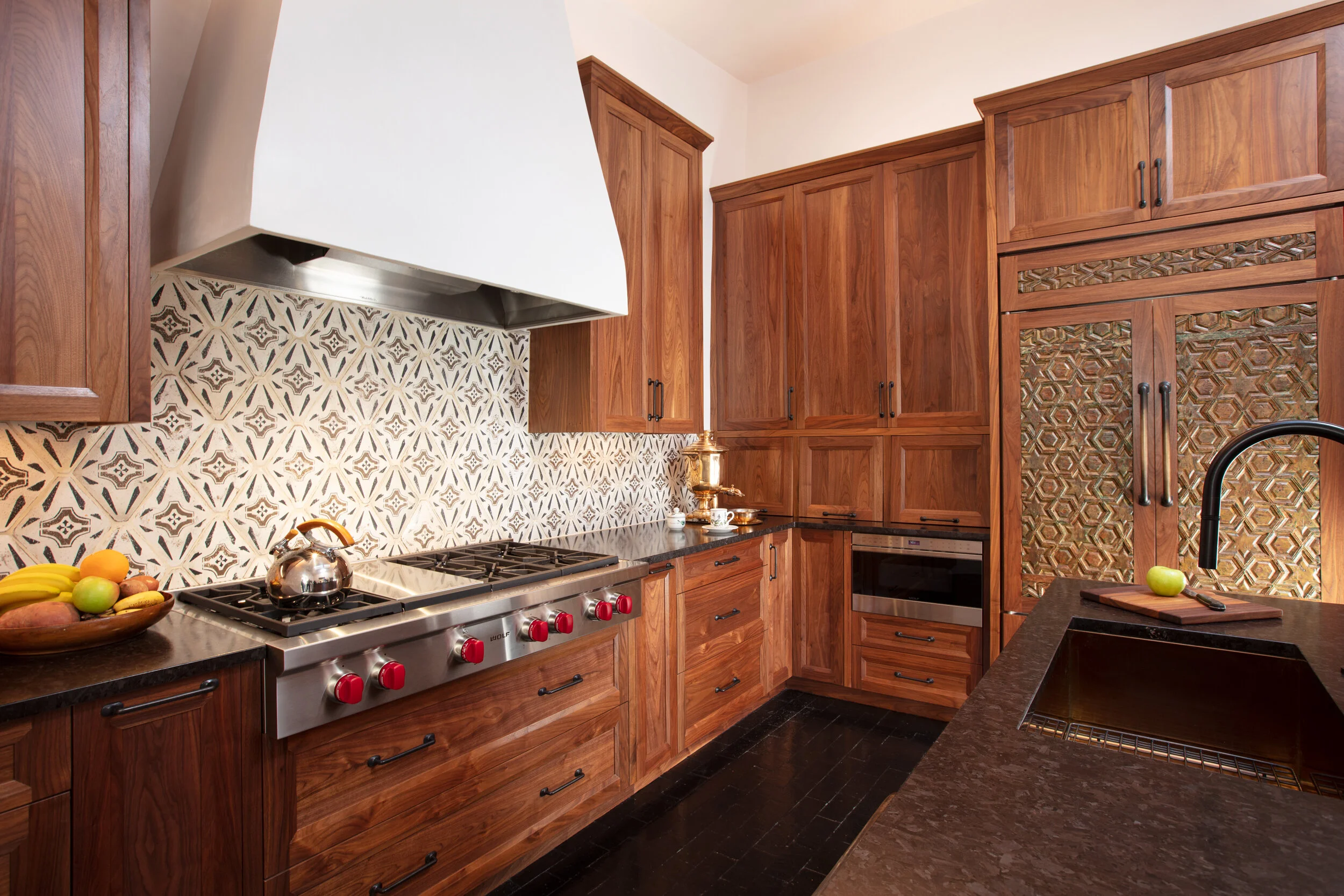 Kitchen with wooden cabinets, black countertops, patterned backsplash, stainless steel stove with red knobs, a bowl of bananas, apples, and oranges, a black faucet, and a cutting board with a green apple.