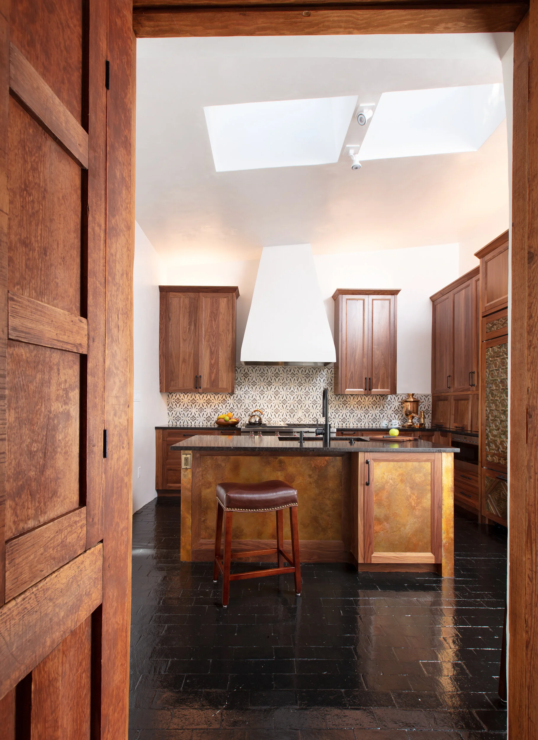 View into a rustic kitchen with wooden cabinets, black countertops, and a patterned tile backsplash. A kitchen island with a leather stool, skylights, and black flooring are also visible.