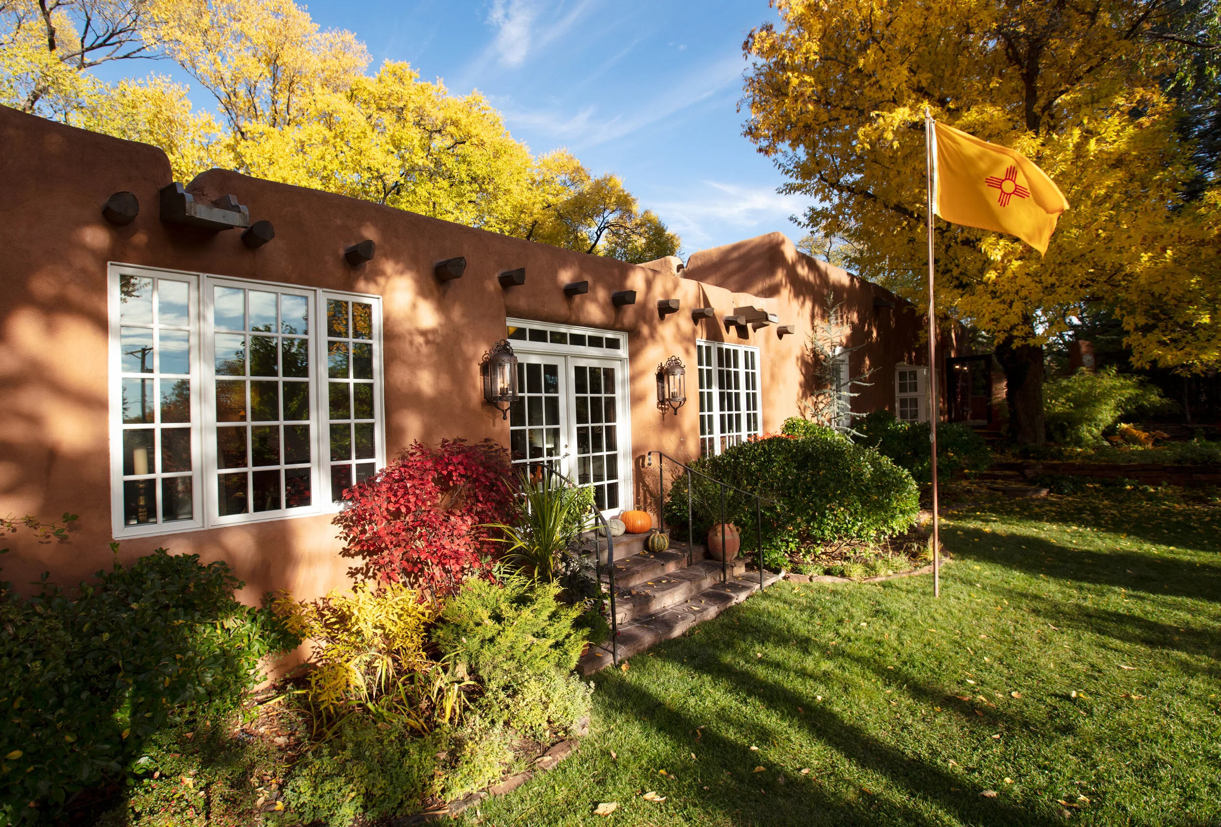 A Southwestern-style house with stucco walls and white-framed windows, surrounded by colorful autumn trees, bushes, and a well-maintained lawn, with pumpkins and autumn decor on the steps leading to the door, and a large yellow flag with a red symbol