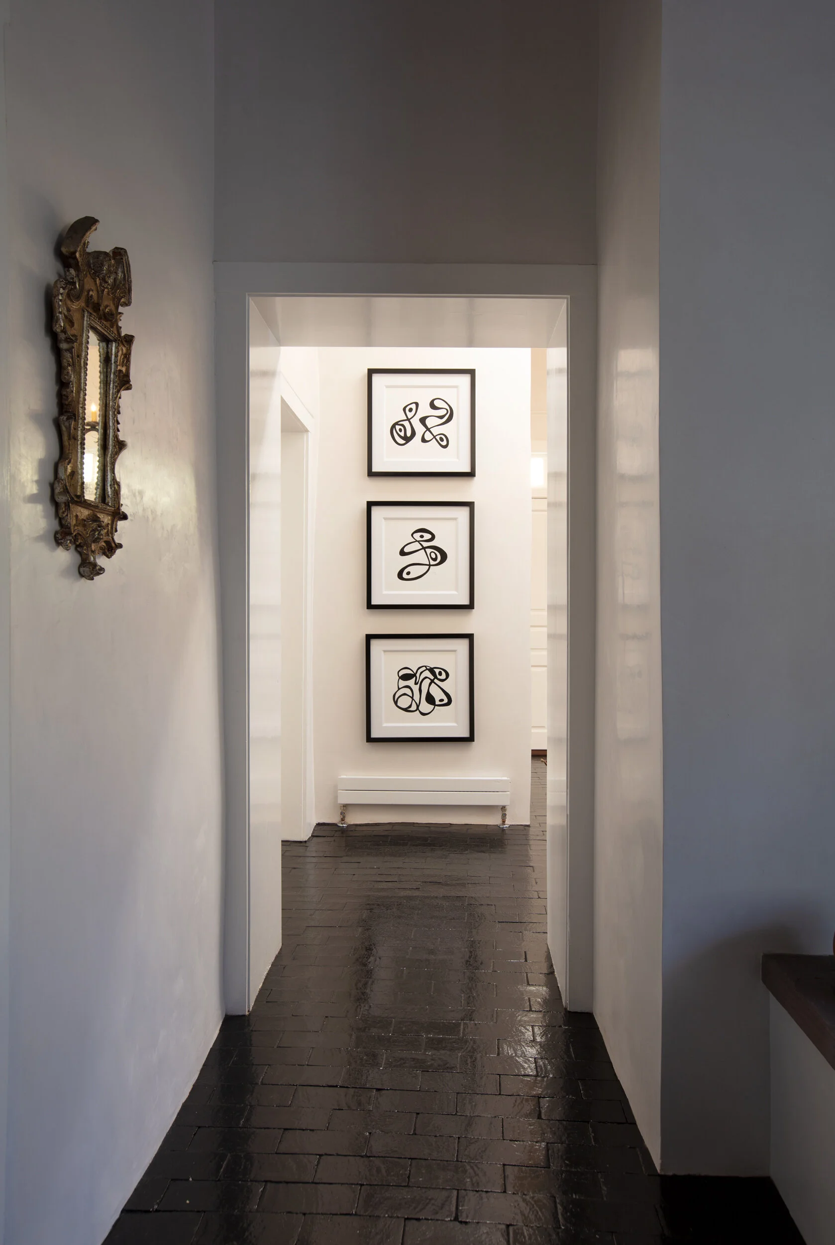 A narrow hallway with black glossy floor tiles, white walls, a decorative gold-framed mirror on the left, and three framed abstract black line art prints on the far wall.