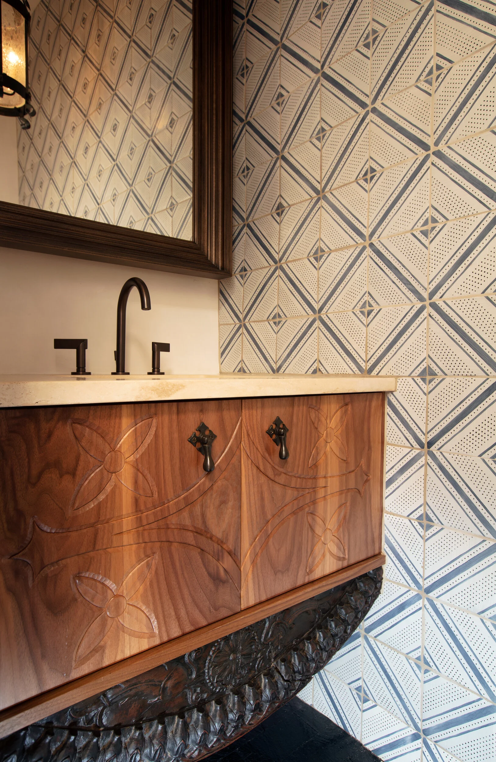 Close-up of a bathroom vanity with a wood cabinet featuring carved floral patterns, a light-colored stone countertop, a black faucet, and a mirror mounted on a wall with decorative blue and white geometric patterned tiles.
