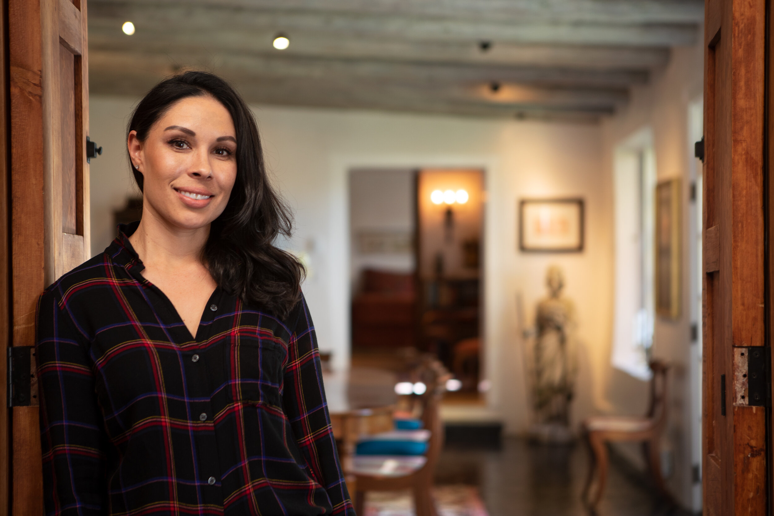 A woman with dark hair and a black plaid shirt smiling in a cozy, well-lit living space with wood accents and framed artwork on the walls.