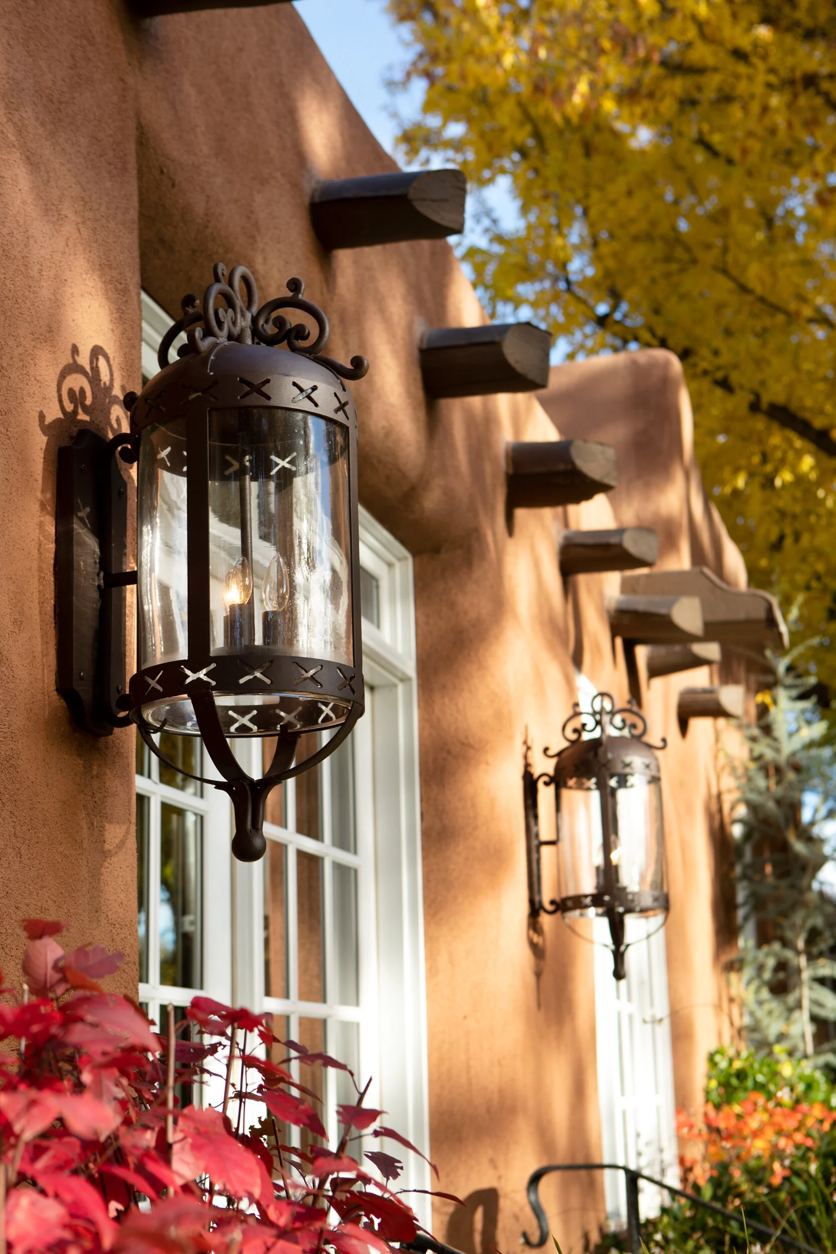 Exterior of a building with a stucco wall, featuring white-framed windows and decorative black metal lanterns, surrounded by colorful autumn foliage.