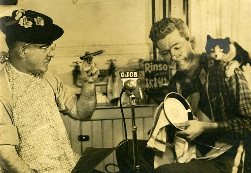 Bill Loeb (smoking), Lloyd MacInnis (washing dishes), and a cat (looking at you) around a CJCB microphone,