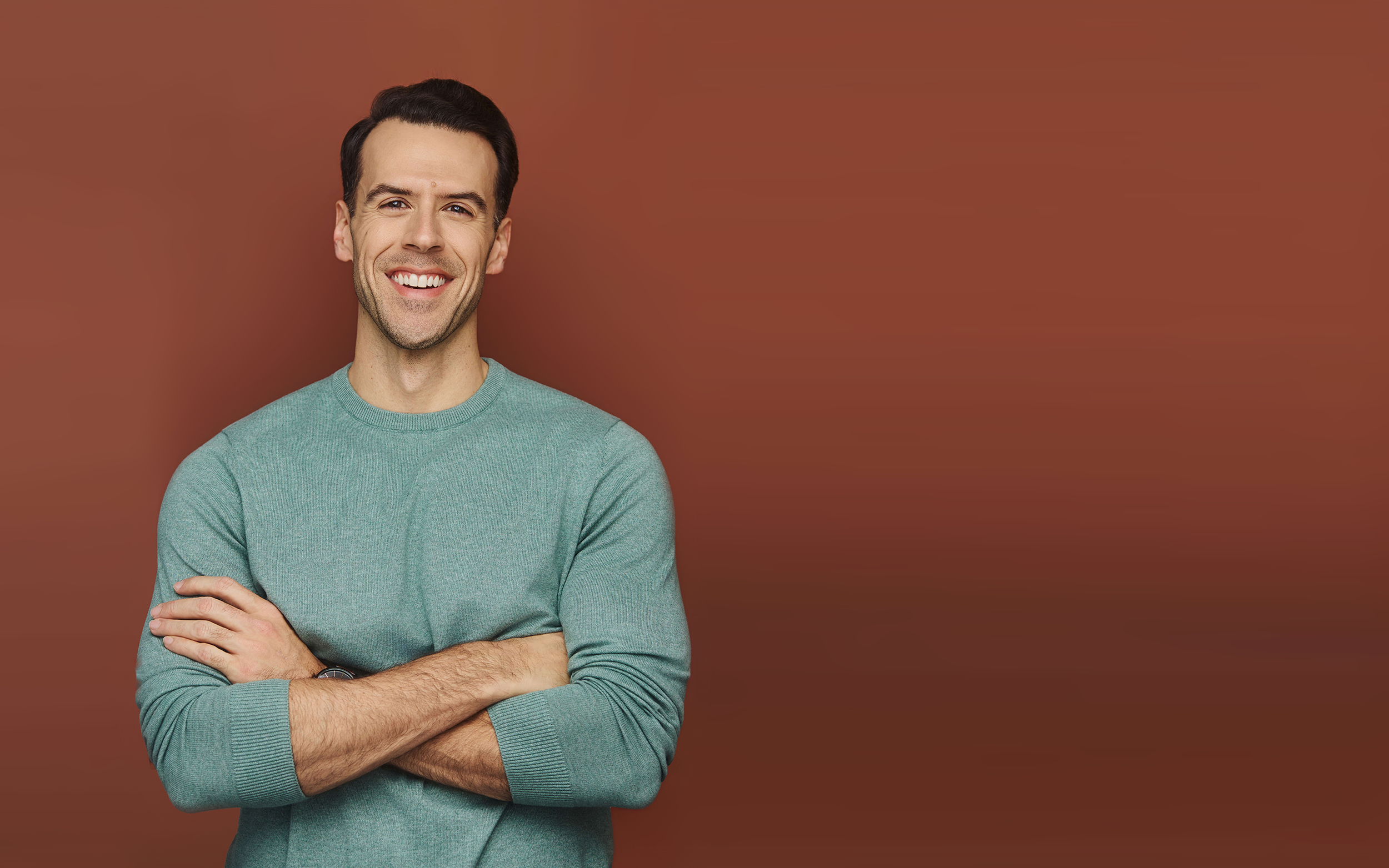 A man with dark hair smiling and crossing his arms, wearing a green sweater, against a brown background.