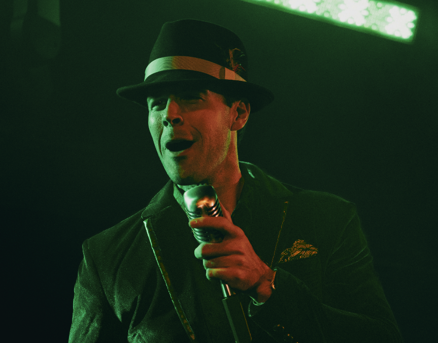 Male stand-up comedian wearing a black fedora with a white band and black blazer, holding a vintage microphone and performing on stage with green lighting.