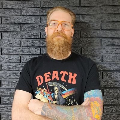 A man with glasses and a beard standing against a black brick wall, wearing a black T-shirt with a graphic of Grim Reaper and colorful tattoos on his arms.