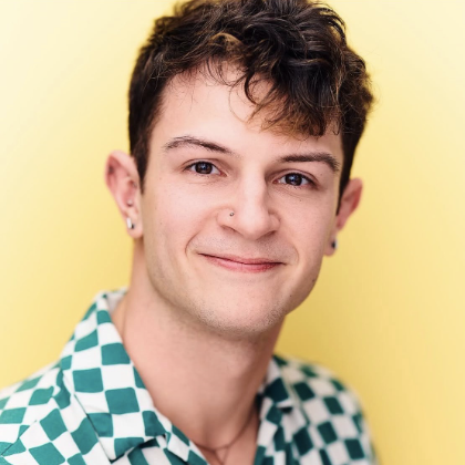Portrait of a young man with short dark hair, wearing a checkered shirt and earrings, smiling against a yellow background.