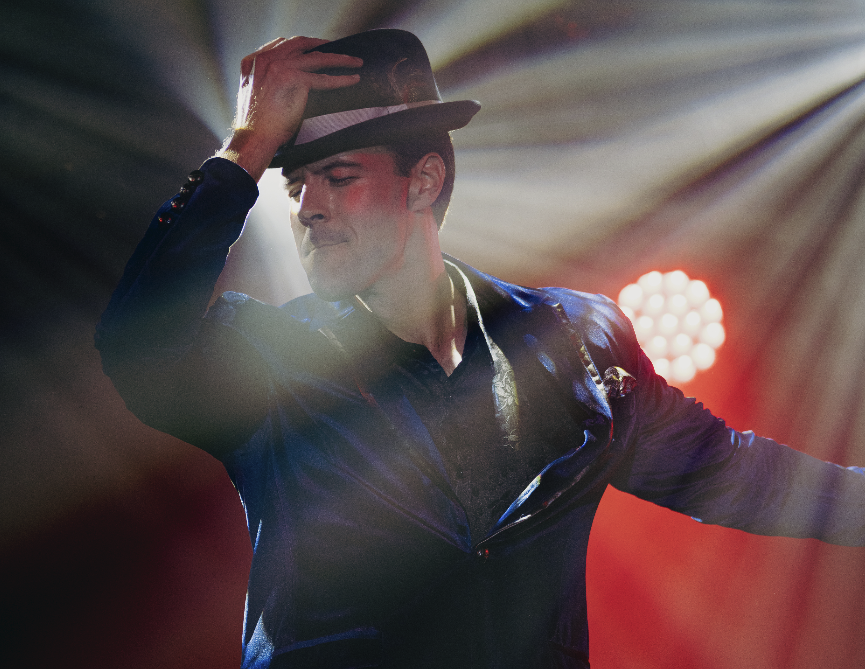 A man wearing a black leather jacket and a hat, holding his hat with one hand, appears to be dancing on stage with colorful lighting.