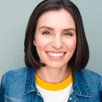 Close-up of a woman smiling with straight, shoulder-length dark hair, wearing a yellow and white shirt with a denim jacket, against a light background.