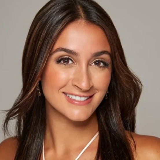 A young woman with long, dark brown hair, smiling with white teeth, wearing subtle makeup and diamond earrings, against a plain grey background.