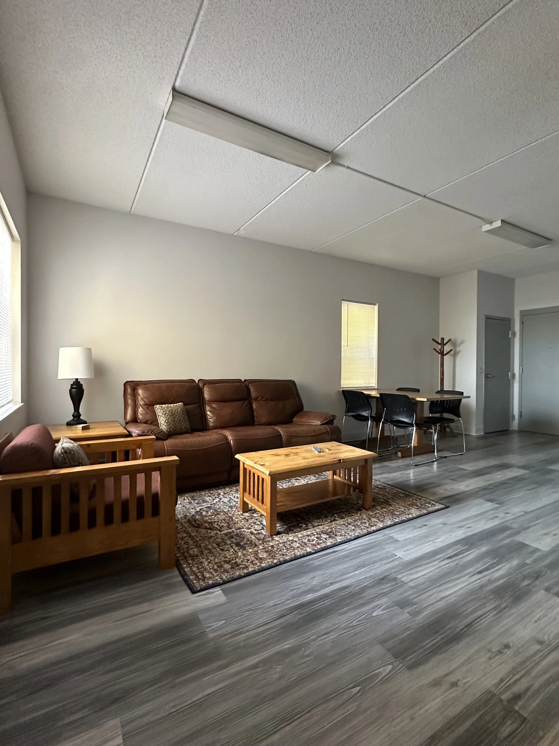 Living room with a brown leather sofa, wooden coffee table, and wooden armchair. There is a patterned rug underneath the coffee table, a lamp on side table beside the sofa, a dining table with black chairs, and a coat rack in the background. The floor is gray wood, and the room has white walls and a ceiling with fluorescent lights.