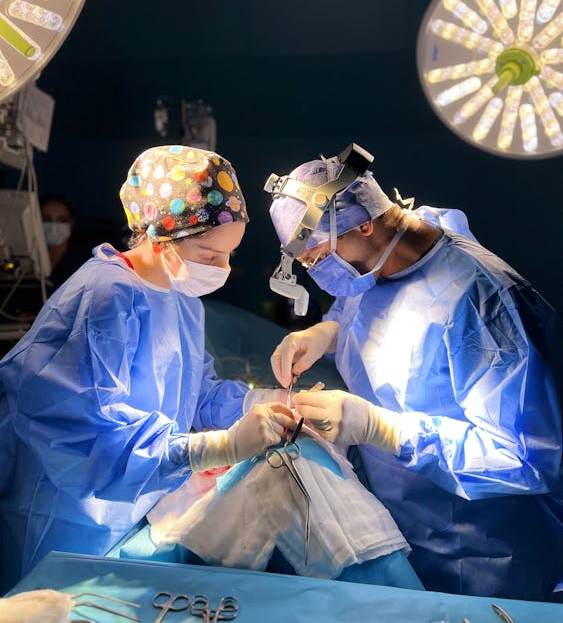 Two surgeons wearing scrubs and masks work under bright surgical lights in an operating room. Available from Stitch Texas medical apparel.