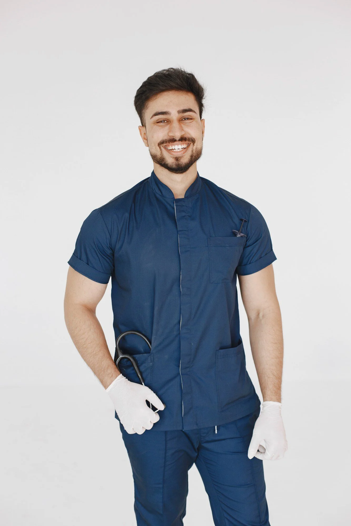 A smiling healthcare professional wearing navy scrubs from Stitch Texas, with gloves and a stethoscope, stands against a clean white background.