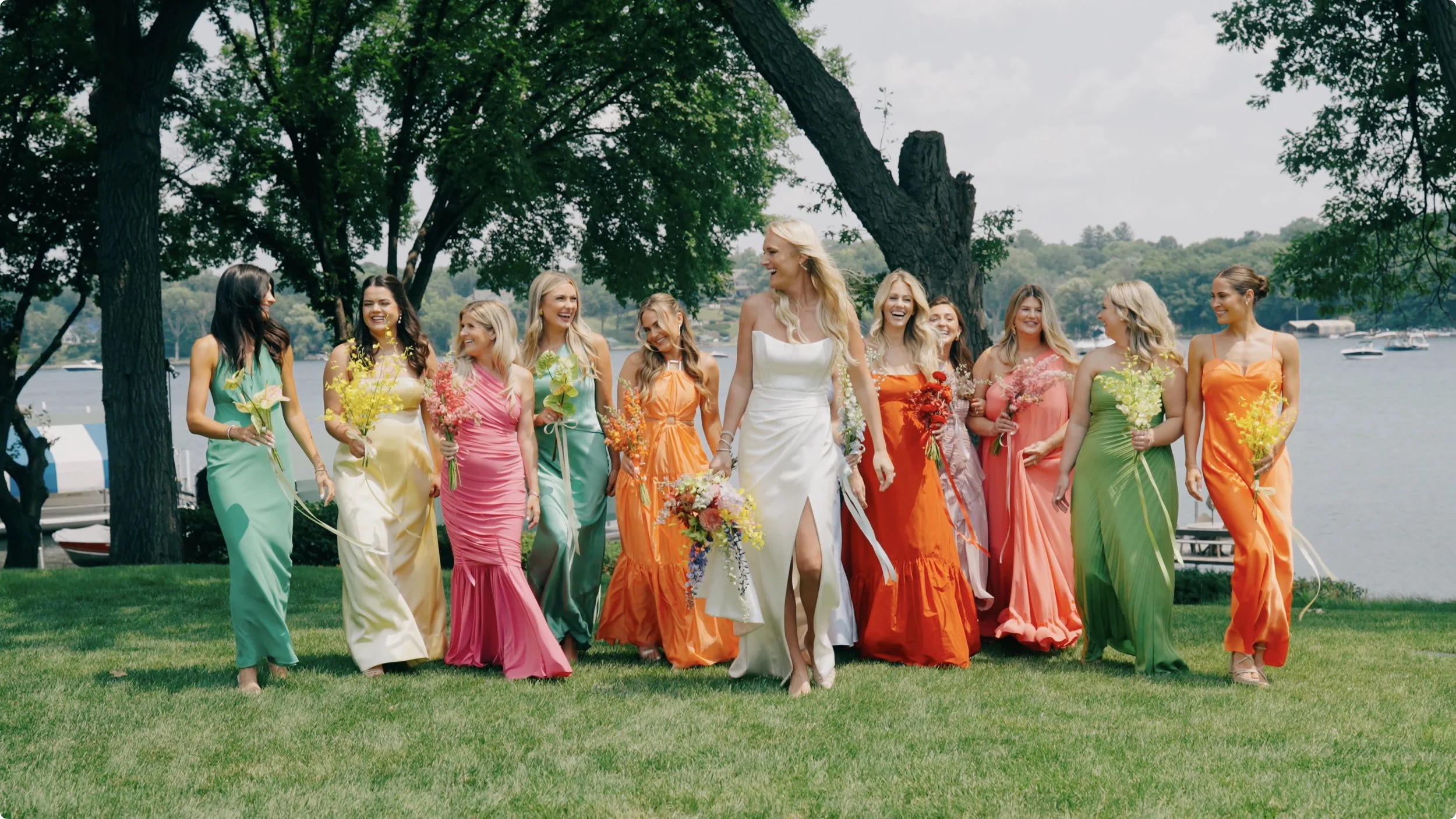 A group of women, including a bride in a white dress, walking on grass near a lake, holding colorful flowers, surrounded by trees.