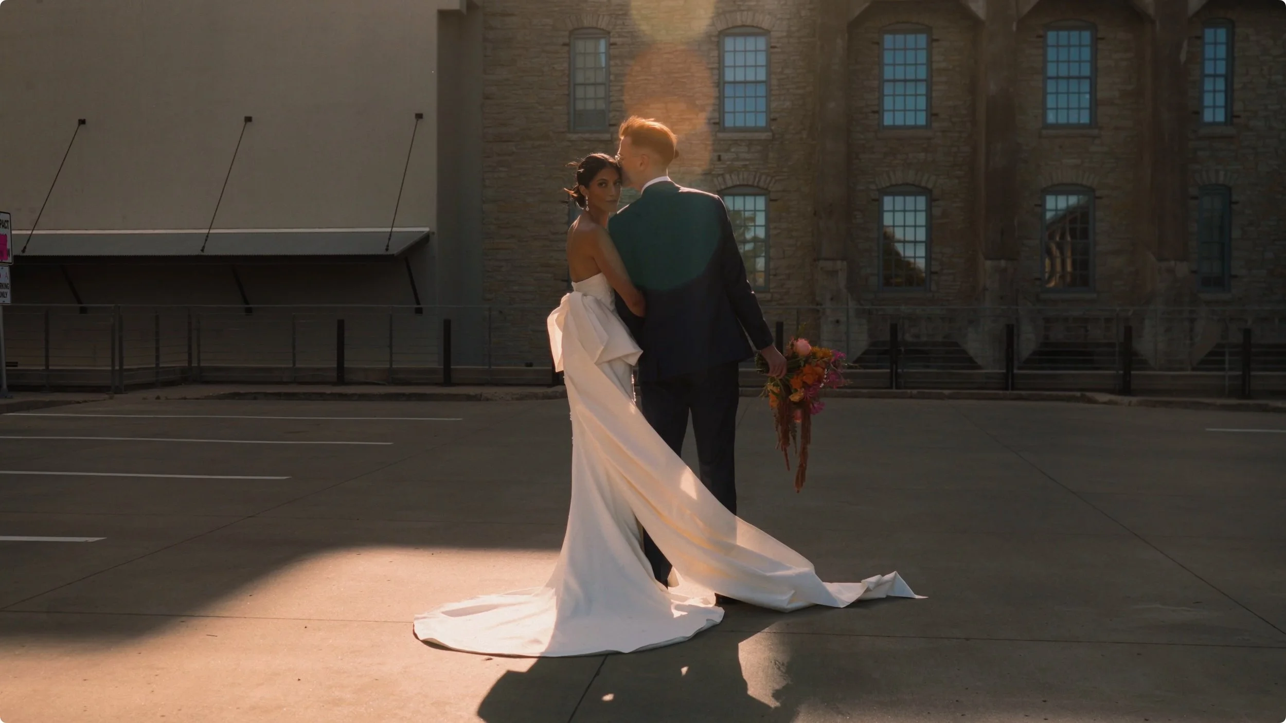 A bride and groom embracing in an empty parking lot during sunset, with a brick building in the background.