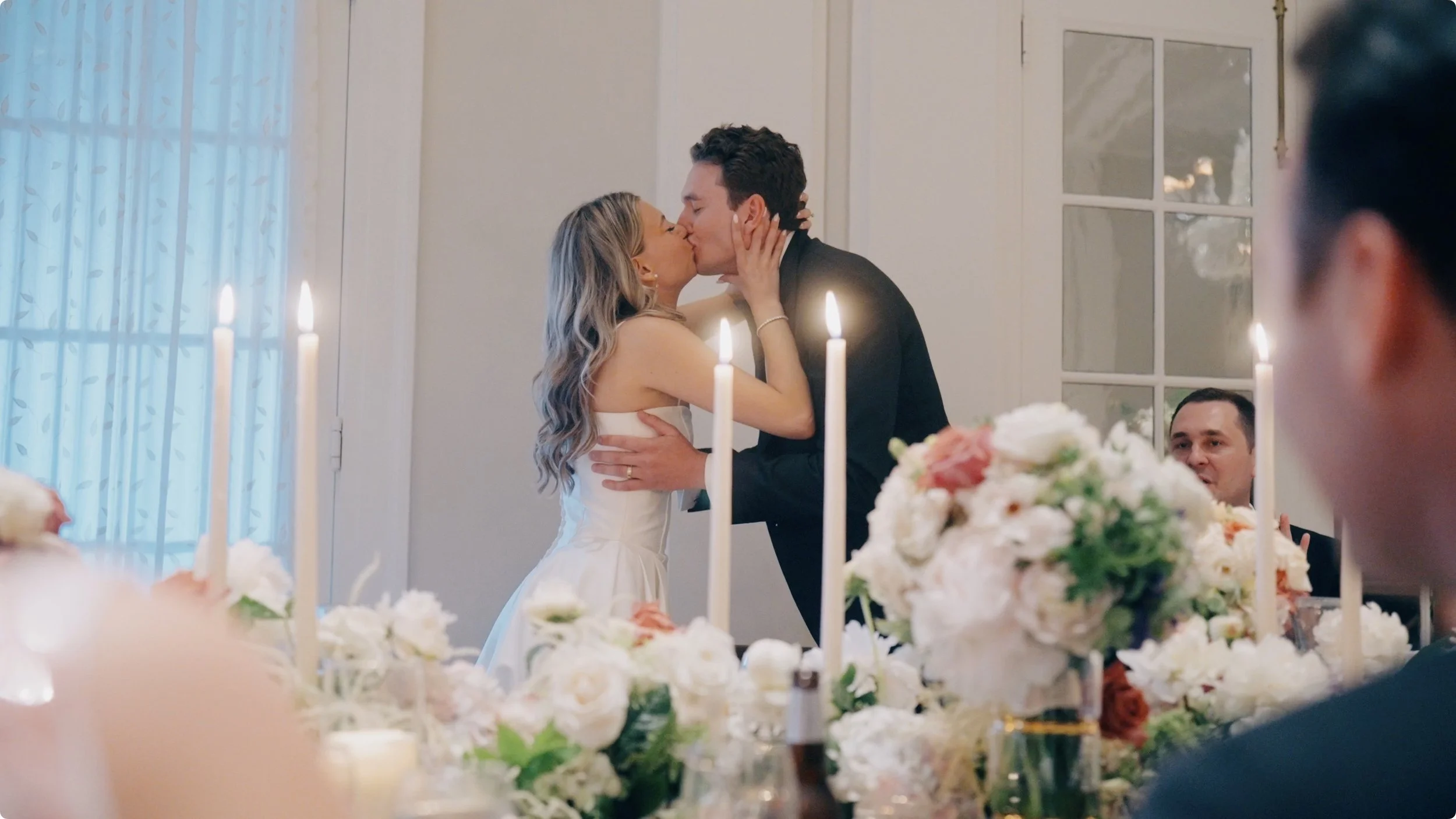 A couple kissing at their wedding reception, surrounded by floral centerpieces and candles, with guests watching and celebrating.