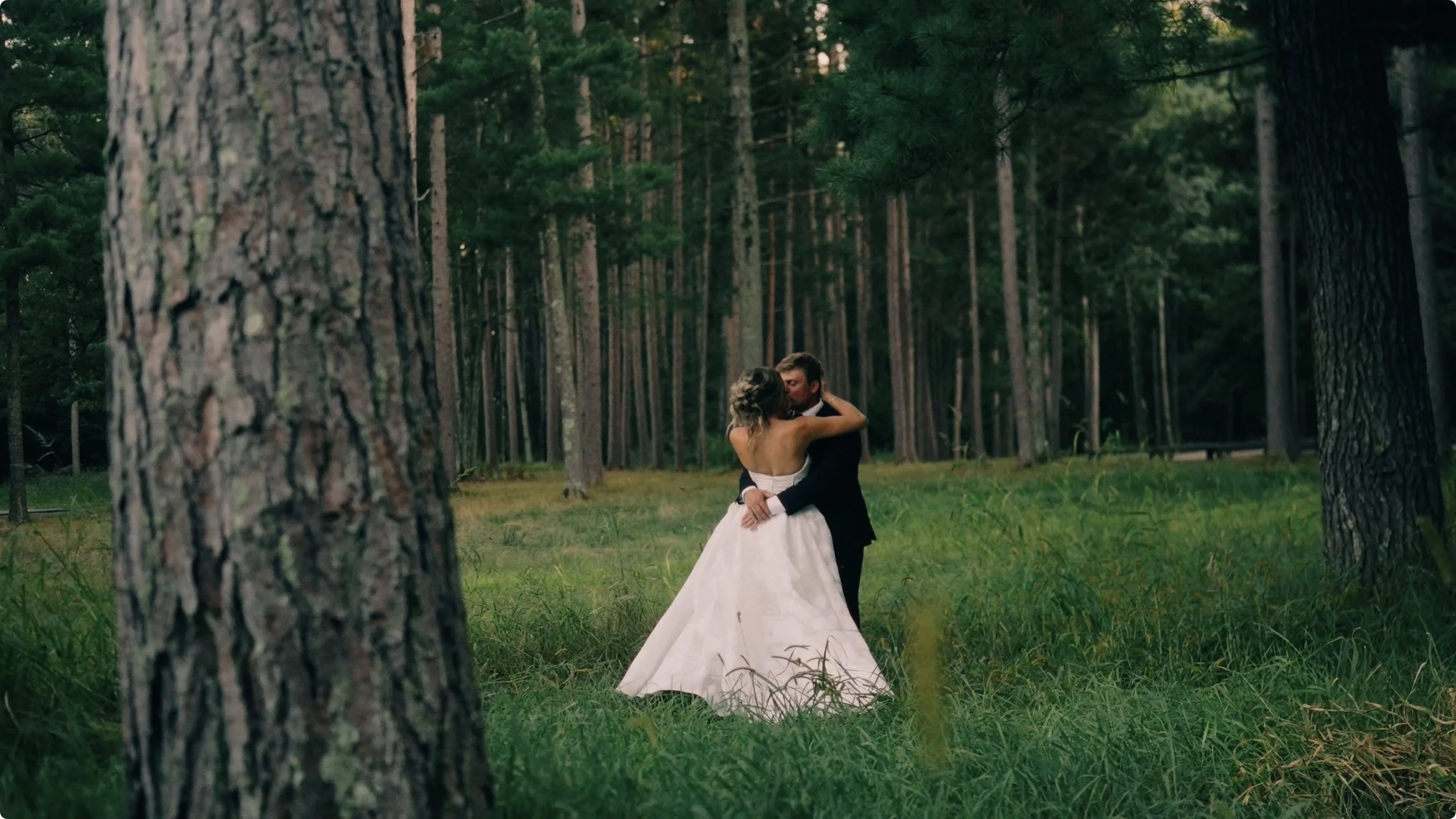 A couple dressed in wedding attire dancing and kissing in a forest clearing surrounded by tall trees and greenery.