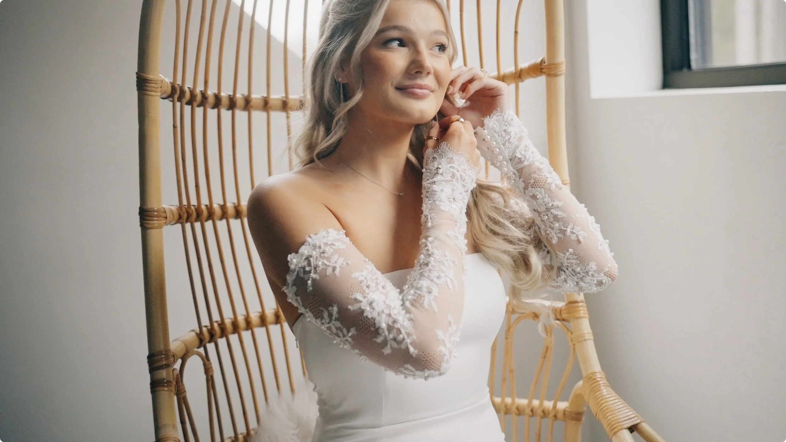 A bride in a white wedding dress with lace sleeves, sitting on a wicker chair near a window, adjusting her earring and smiling.