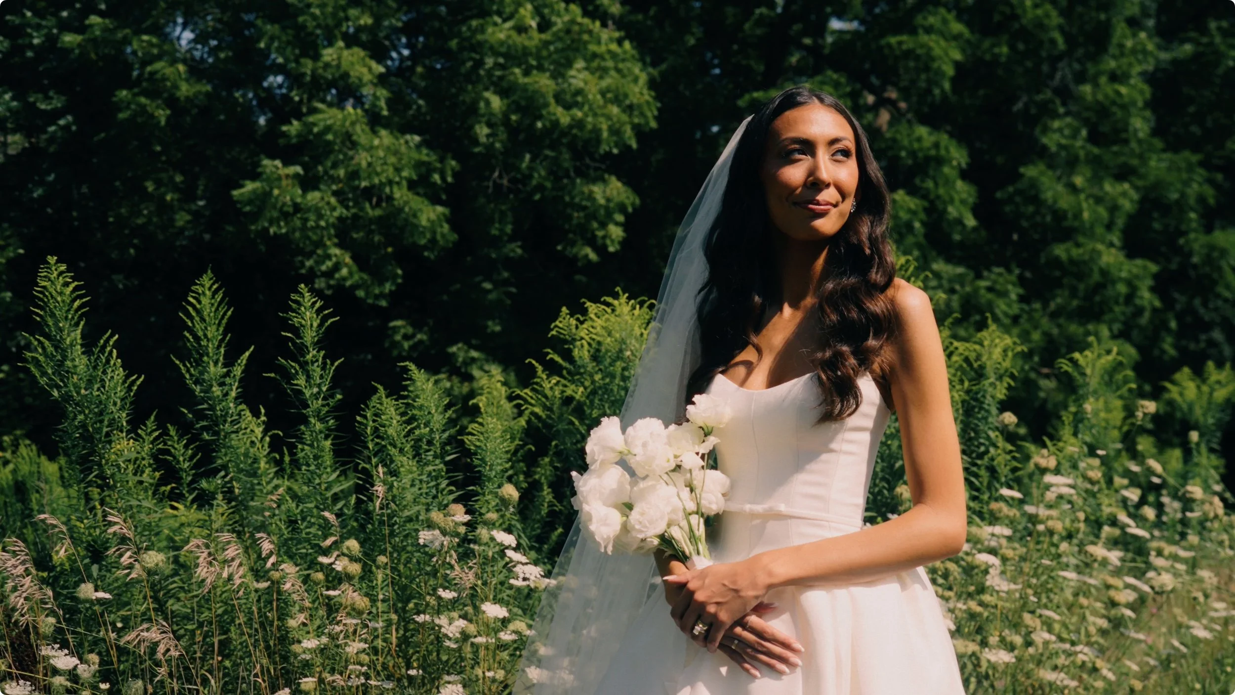 A bride wearing a white wedding gown and veil, holding a bouquet of white flowers, standing outdoors in a lush green garden.